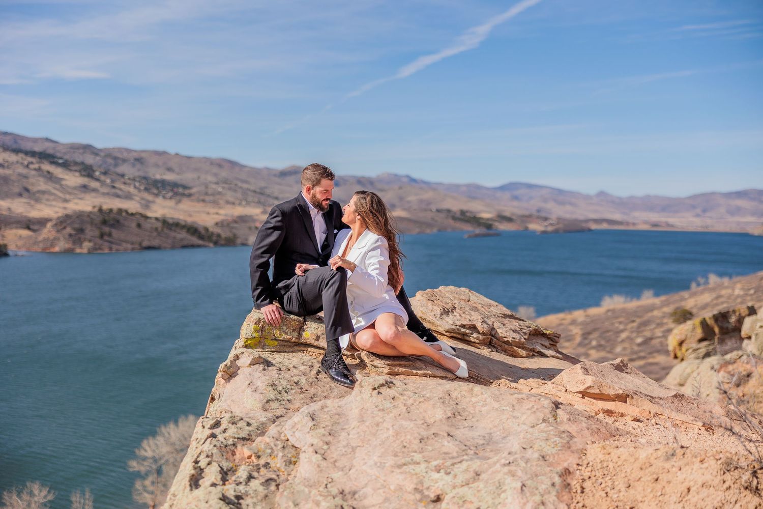 A couple sits on dramatic rocky cliffs overlooking a blue mountain lake.
