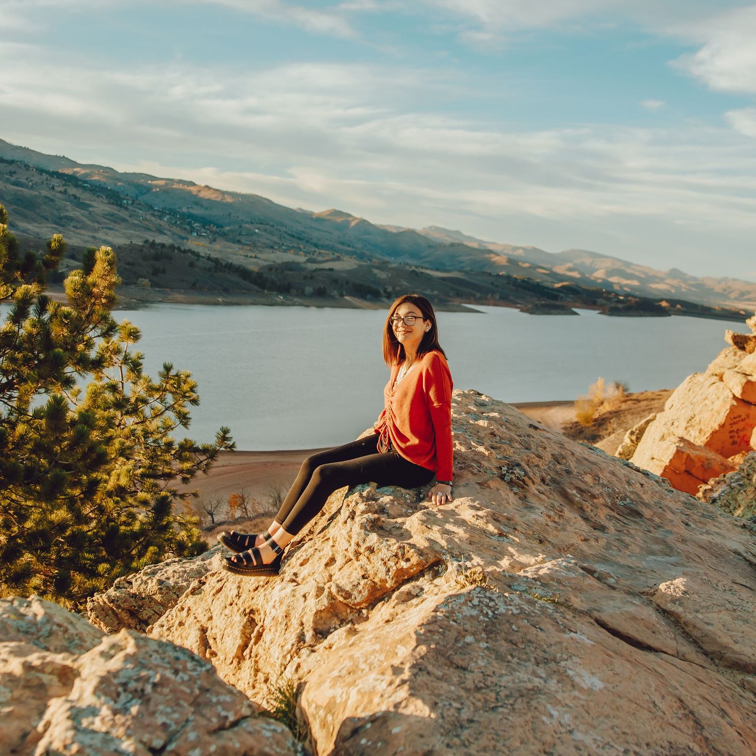 Person in red sweater sits on rocky cliff overlooking scenic mountain lake landscape at sunset.