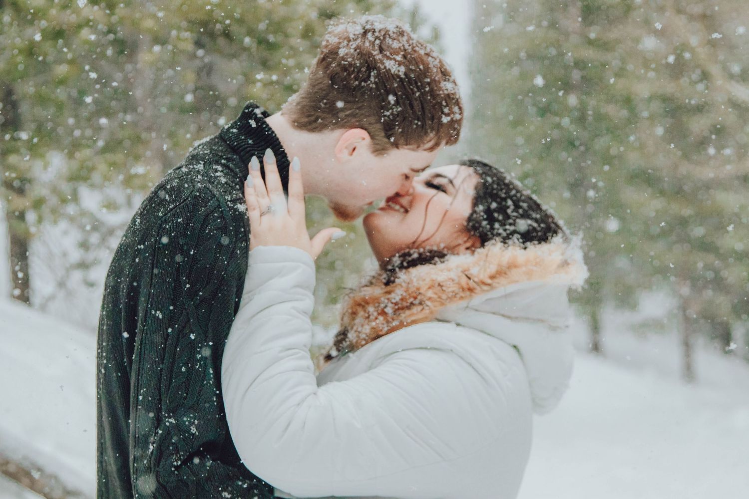 A romantic winter wedding scene with a couple embracing in the falling snow surrounded by evergreen trees.