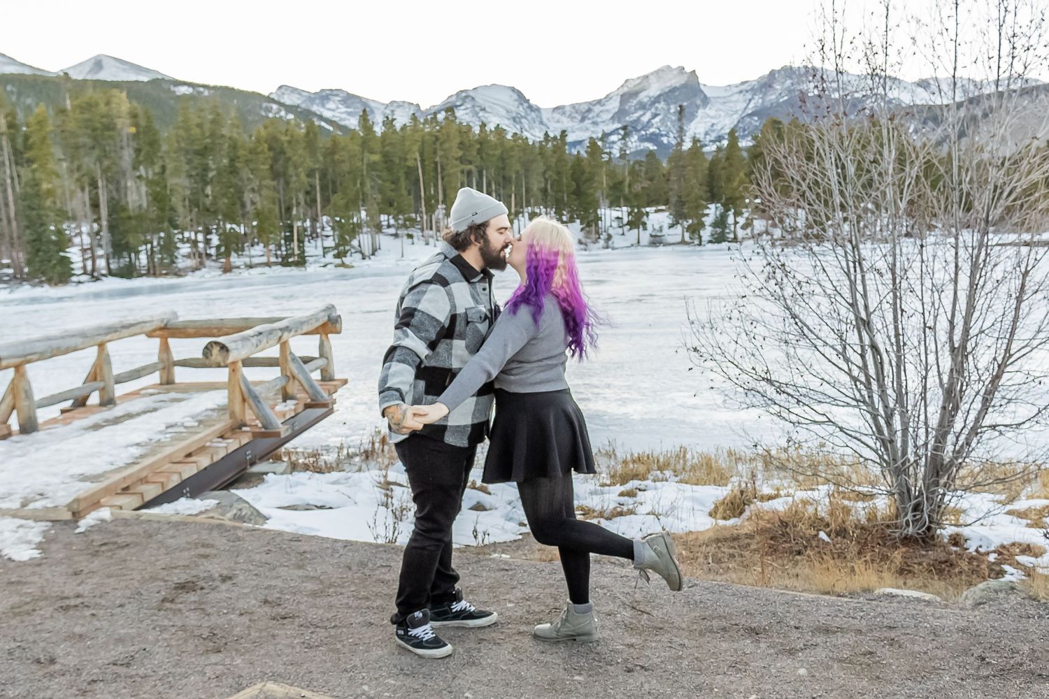Two people share a romantic moment in winter against a snowy mountain backdrop at a scenic overlook.