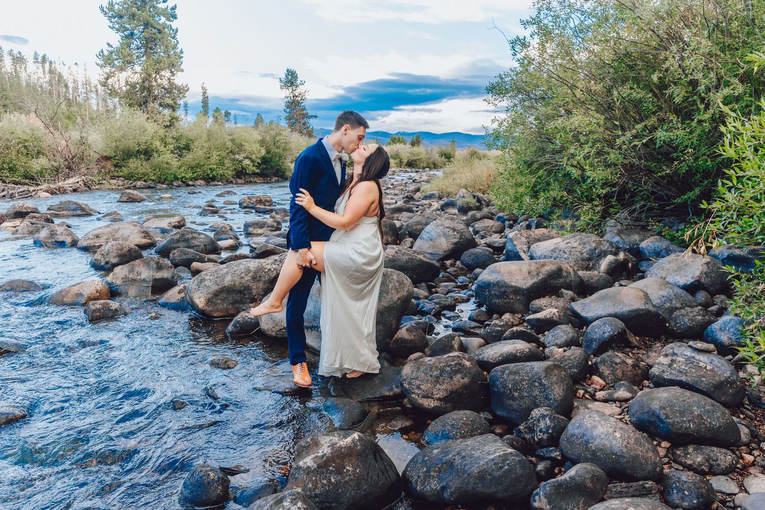 A couple embraces while standing on rocky terrain beside a flowing mountain stream with scenic views in the background.