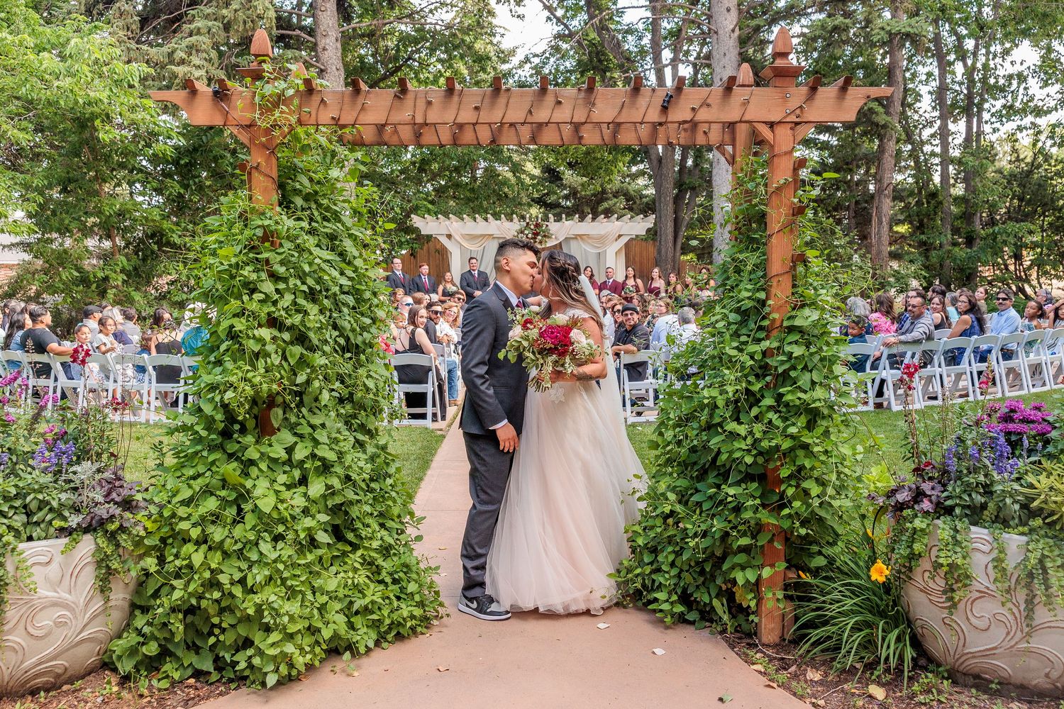 Wedding ceremony setup in a lush garden with wooden pergola and white chairs arranged for guests.