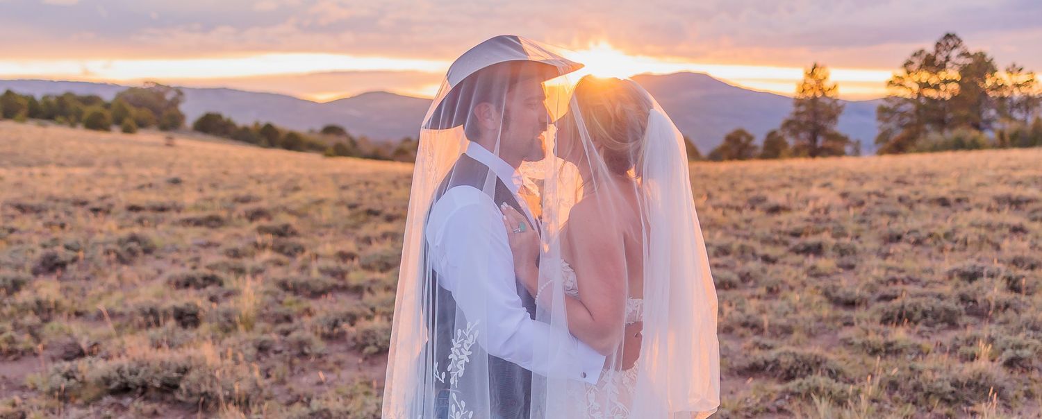 A couple shares a romantic moment at sunset in a field with mountains and golden light streaming through their veil.