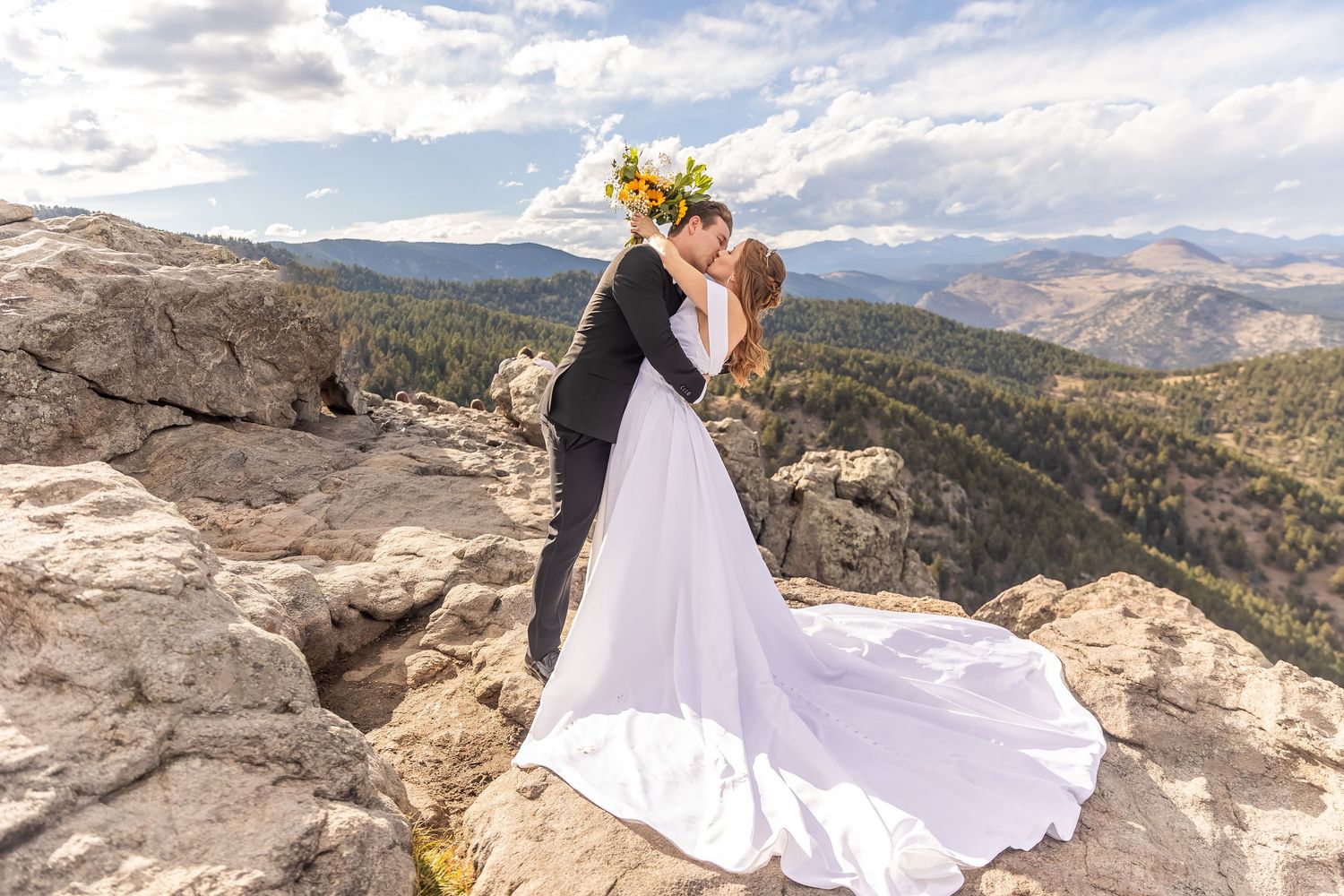 A couple in wedding attire poses on a scenic mountain overlook with dramatic rock formations and sweeping valley views.