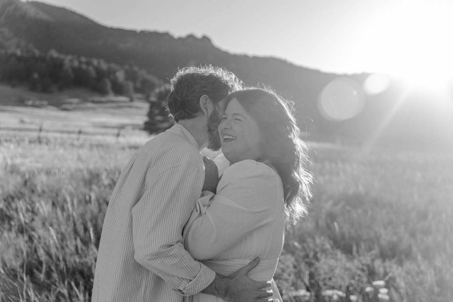 A couple embraces in a sunlit field during golden hour while surrounded by tall grass and mountains.