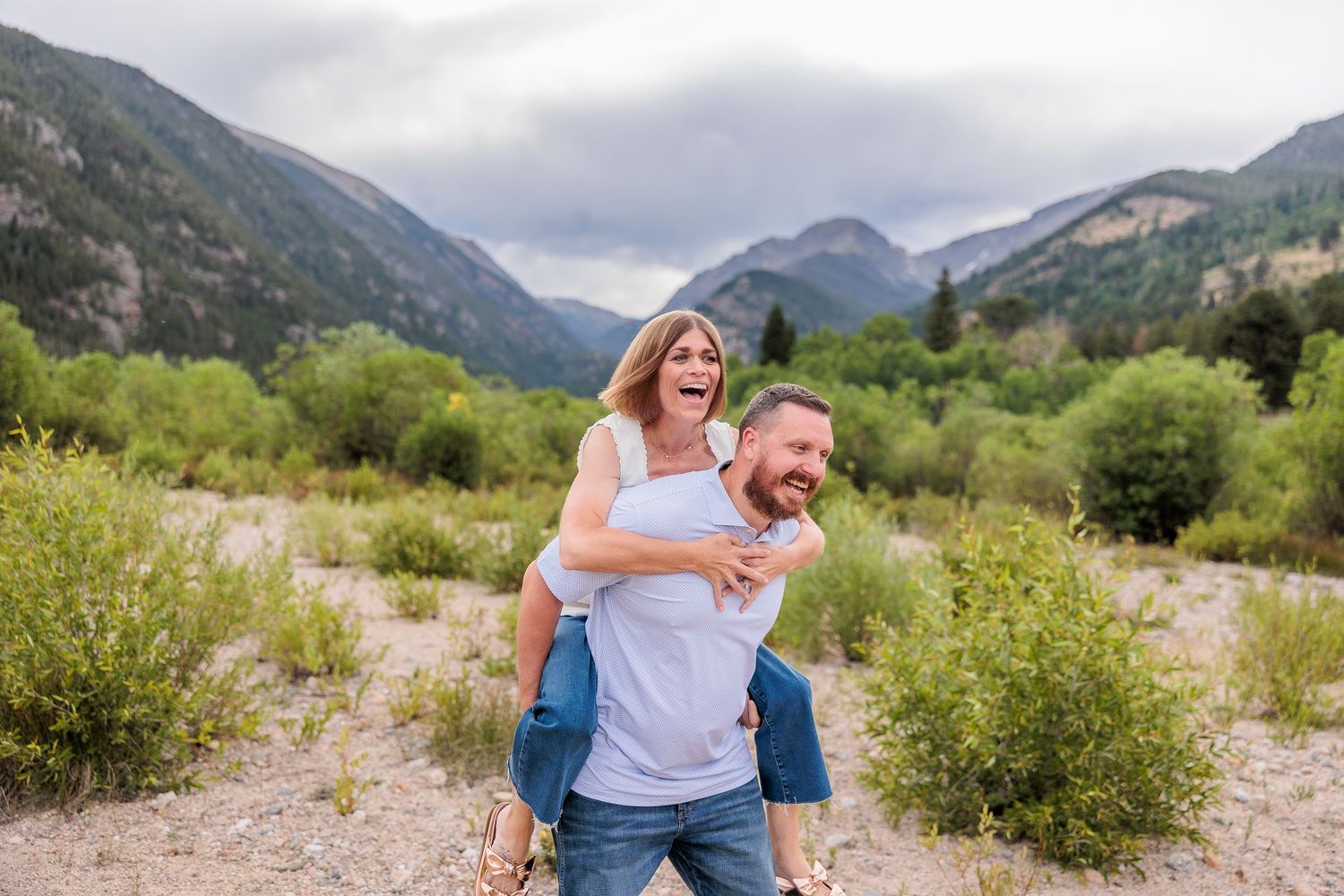 A joyful couple shares a piggyback ride moment in a scenic mountain valley surrounded by green vegetation.