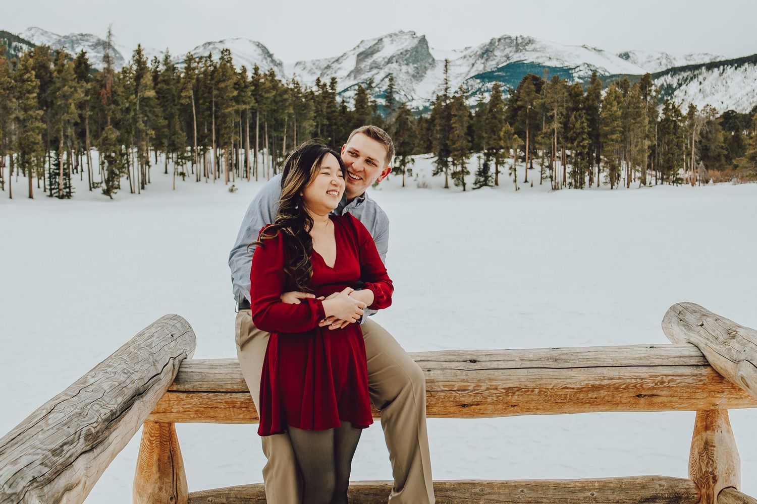 A couple in burgundy and beige attire embraces in a snowy mountain landscape with wooden benches.