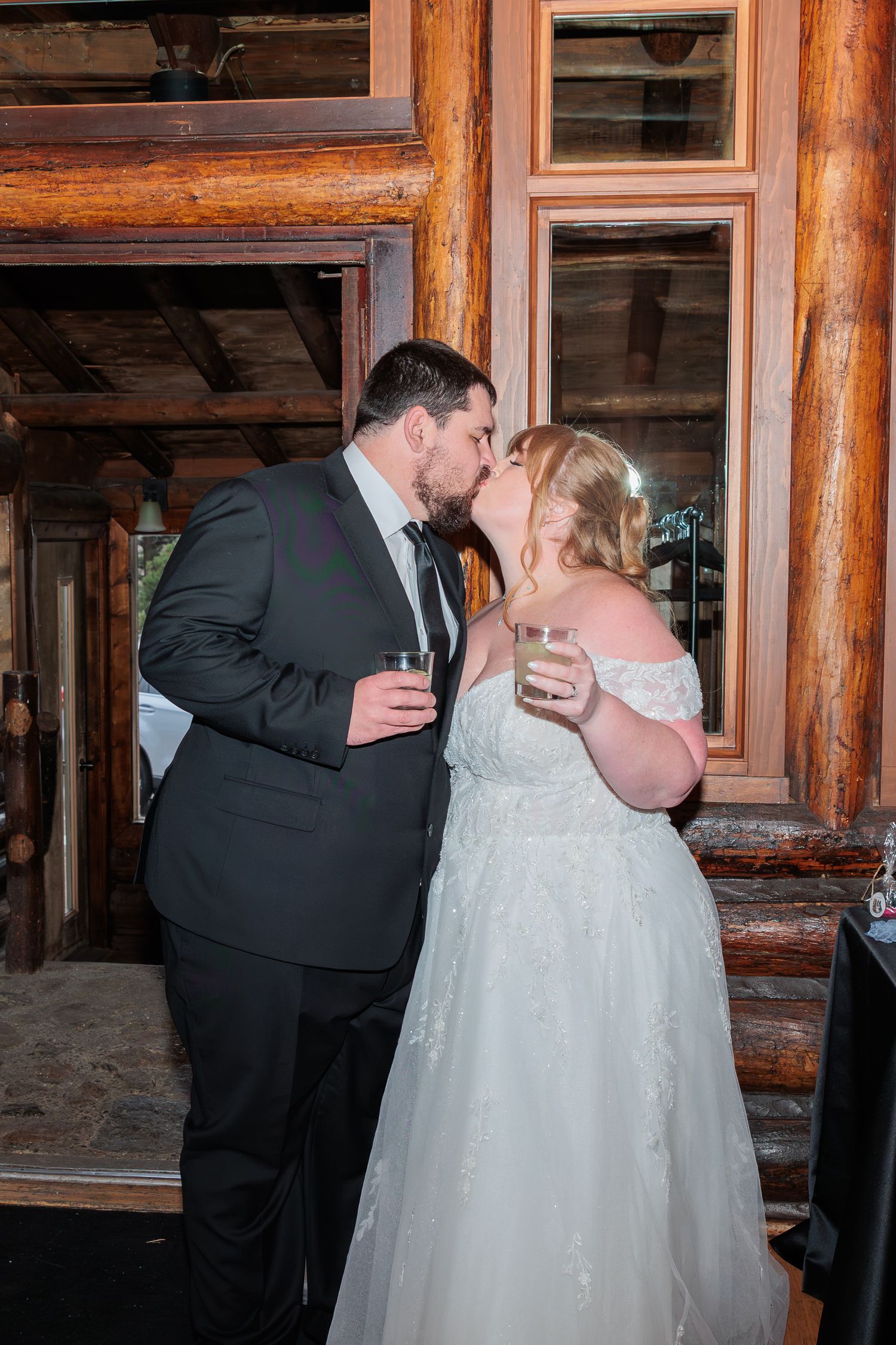 Bride and groom sharing a kiss while holding drinks at a rustic log cabin wedding reception venue.