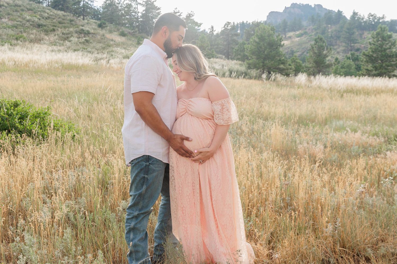 A couple stands together in a golden field during sunset, with the woman wearing a flowing peach-colored dress.