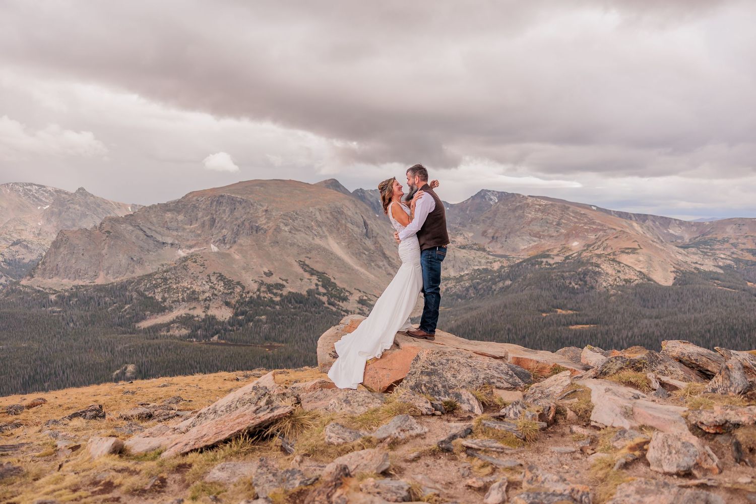 A couple in wedding attire shares a romantic moment atop a scenic mountain vista at sunset.