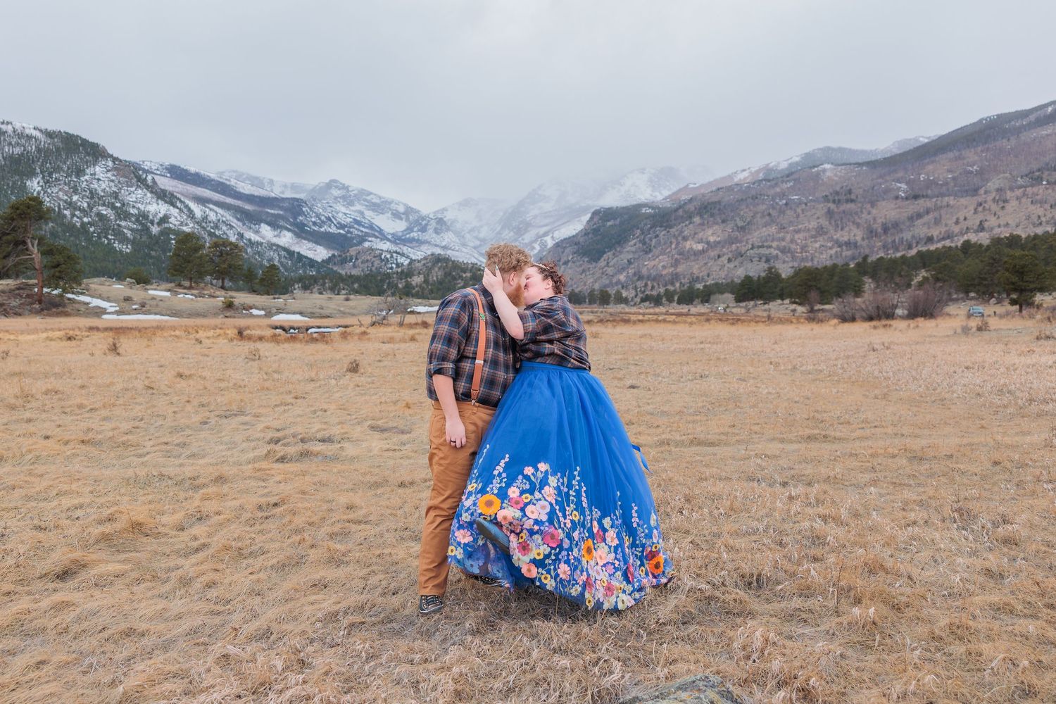 A couple in formal attire poses in a scenic mountain meadow at sunset with snow-capped peaks in the background.