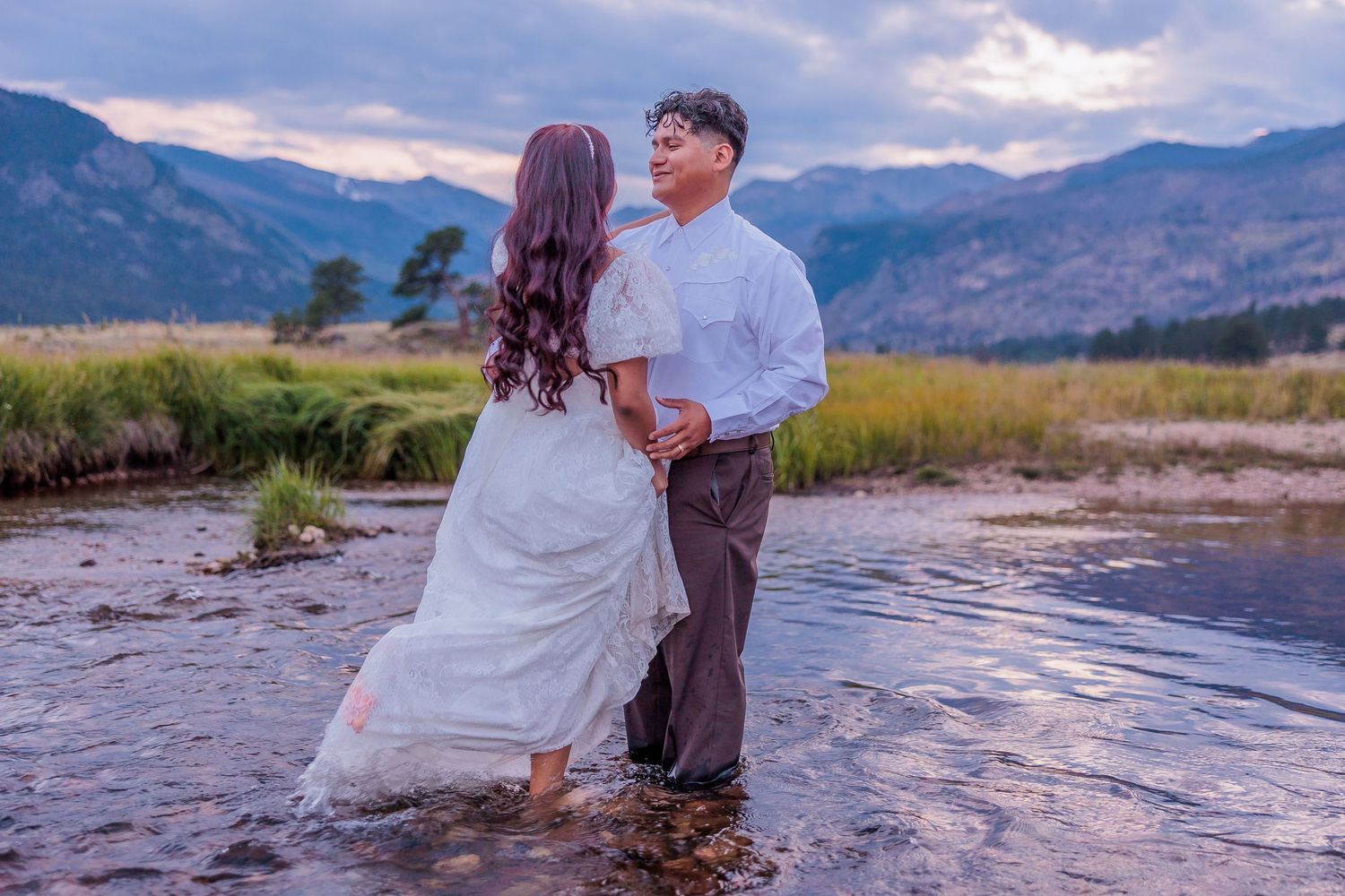 A couple stands in a mountain stream during sunset with mountains and dramatic clouds in the background.