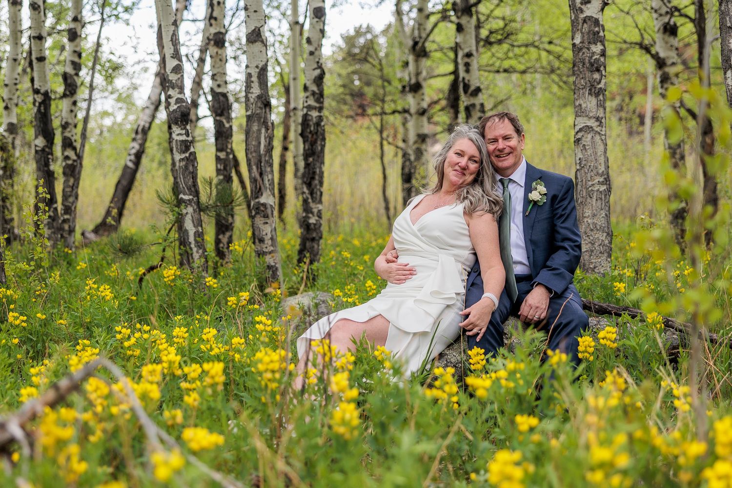 A couple sits together among white birch trees and bright yellow wildflowers in a romantic outdoor setting.