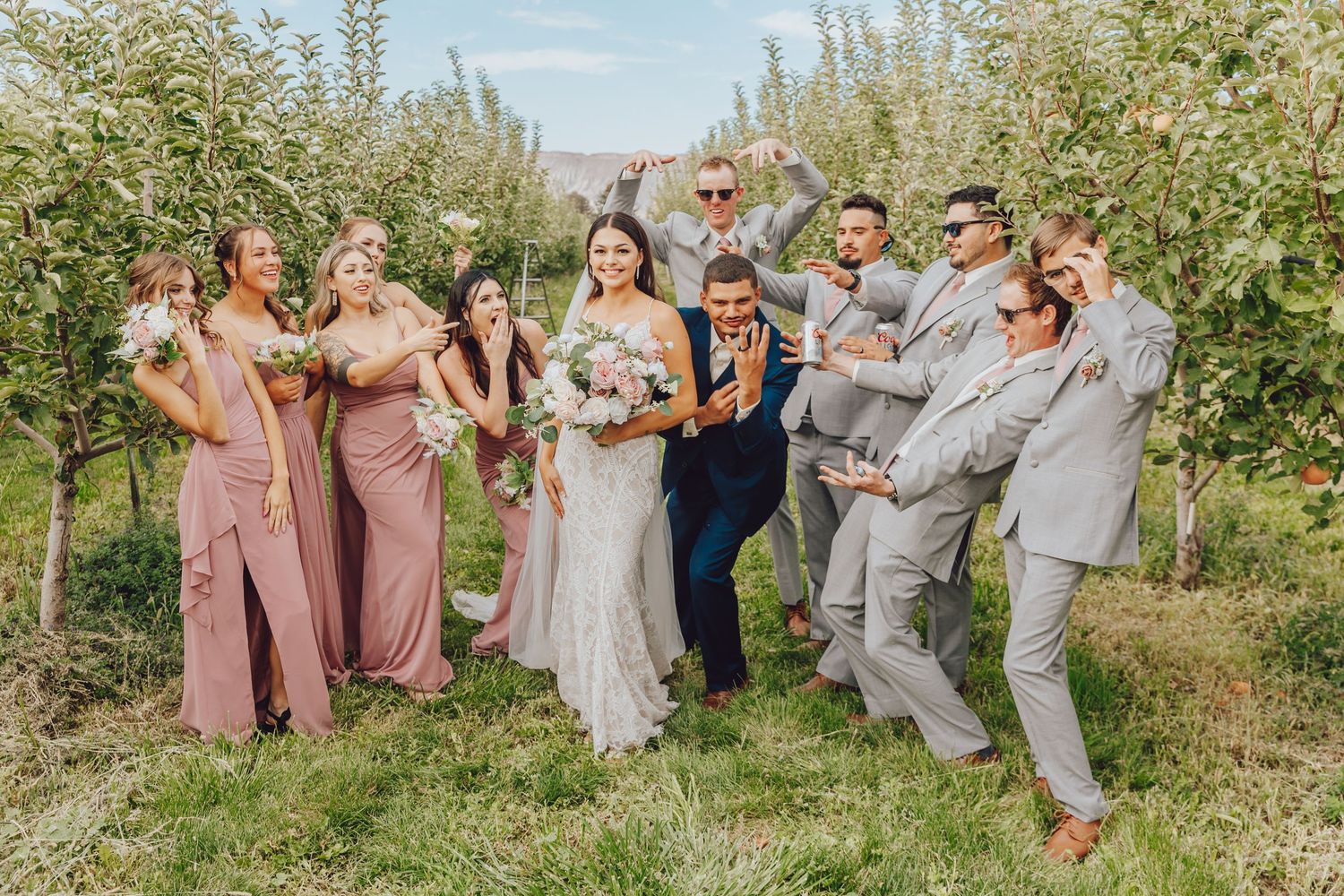 A wedding party poses playfully in an orchard wearing coordinated pink and gray attire.