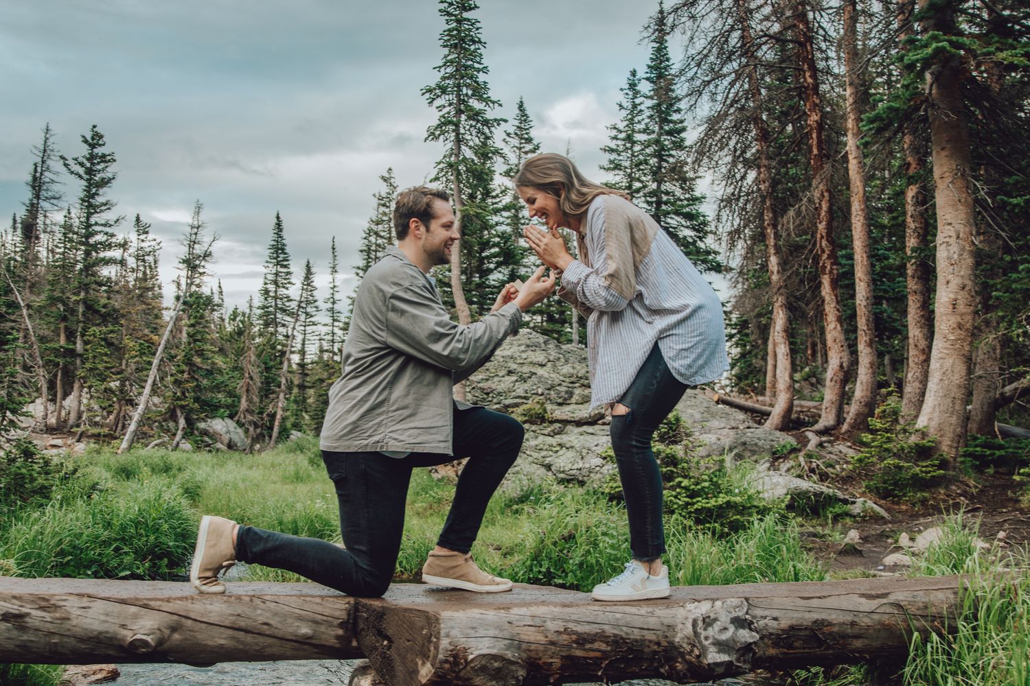 Man proposing to woman on a log in a mountain forest setting, presenting an engagement ring outdoors.