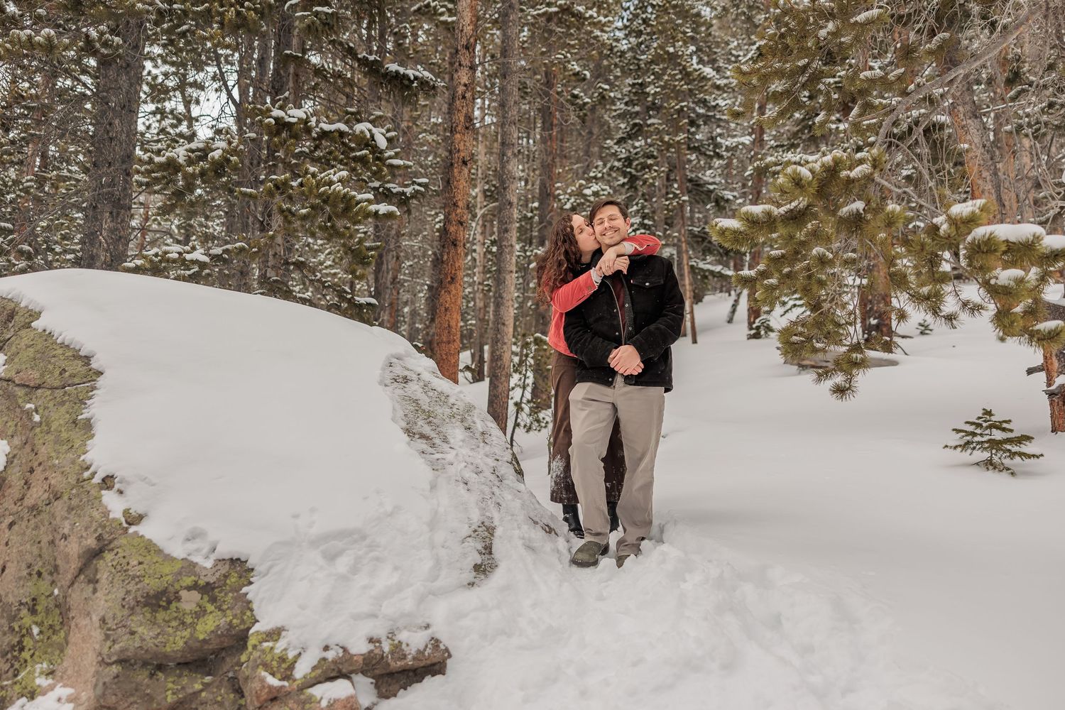 A couple embraces on a snowy winter trail surrounded by evergreen trees in a forest setting.