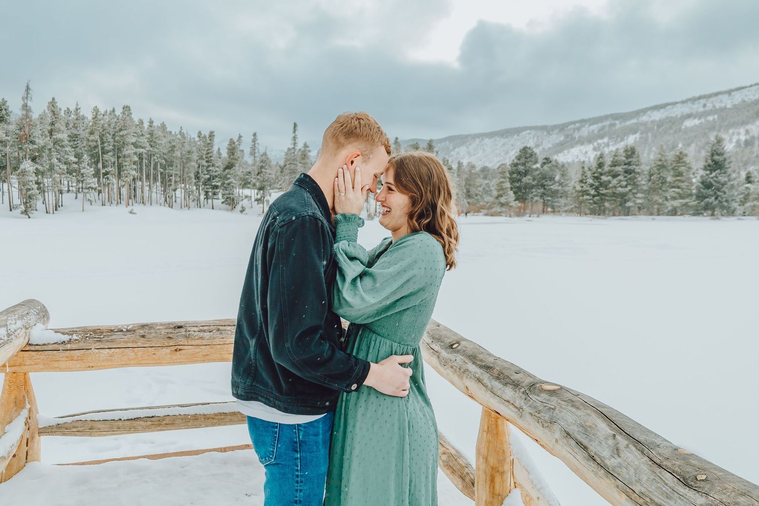 A romantic couple embraces on a snowy mountain overlook with evergreen trees and cloudy skies in the background.