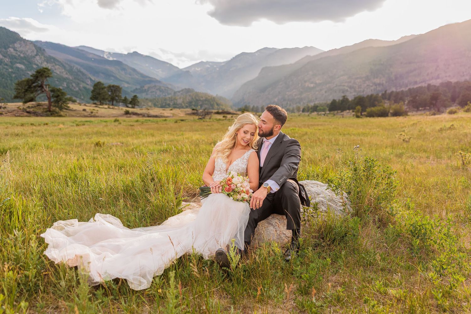 Bride and groom sit on a rock in a lush mountain meadow, with scenic Rocky Mountain peaks in the background.