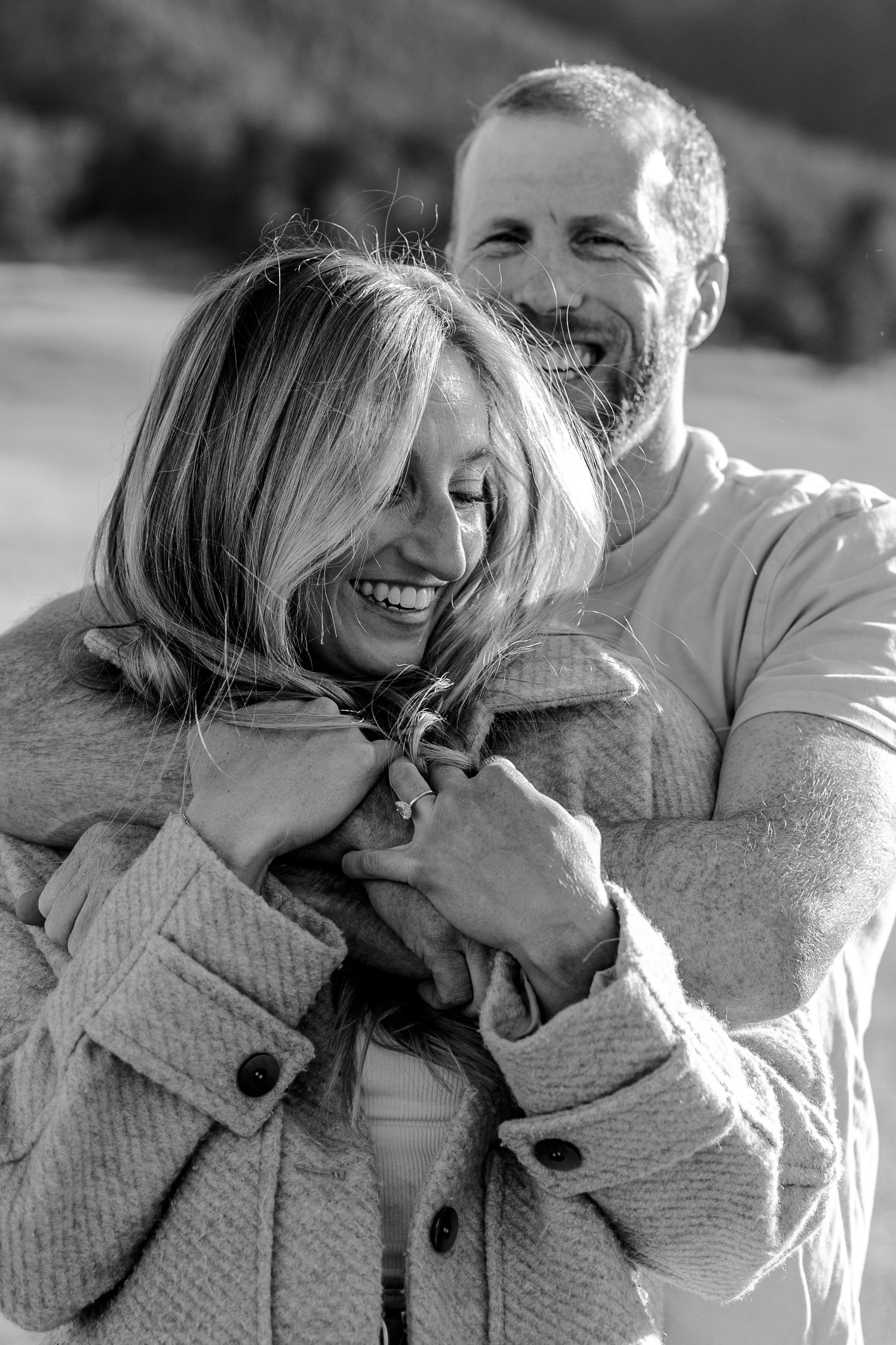 Black and white photo of a smiling couple embracing outdoors, man hugging woman from behind in cozy coats.
