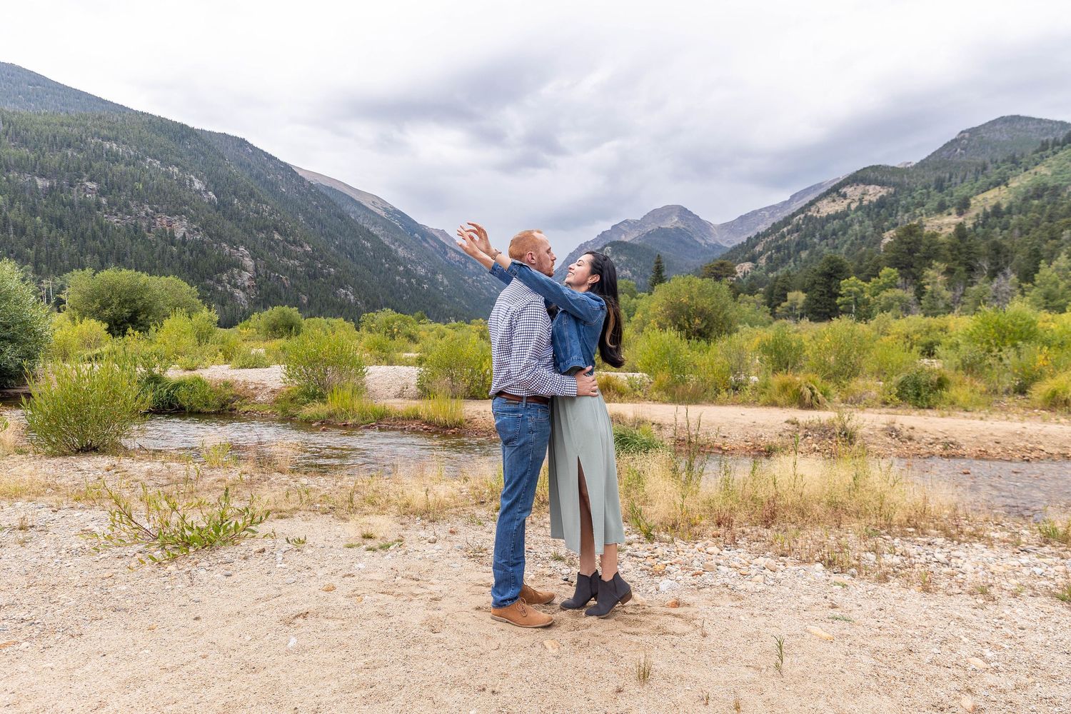 Two people embrace while enjoying scenic mountain views in a grassy valley on a cloudy day.