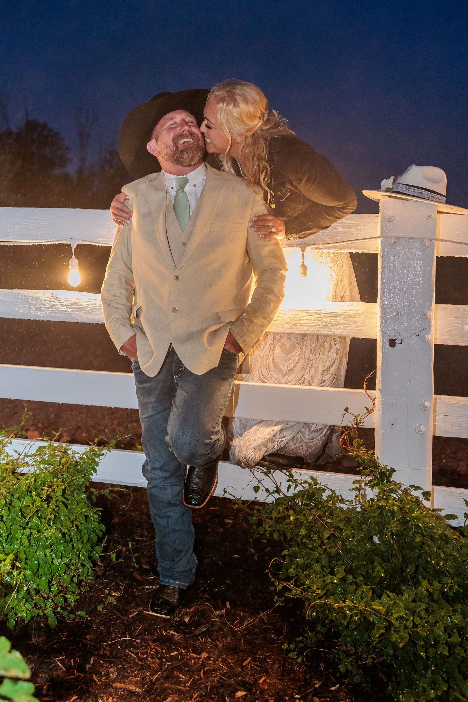 Couple in formal attire leaning against white fence at dusk with lantern lighting and garden foliage below at Rockin S Ranch