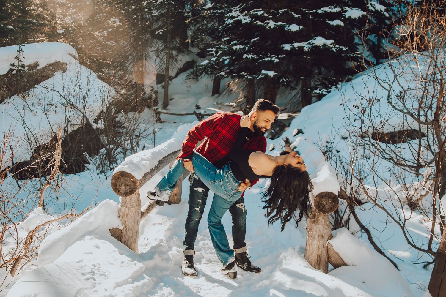 A couple dips romantically in the snow surrounded by evergreen trees on a winter day.