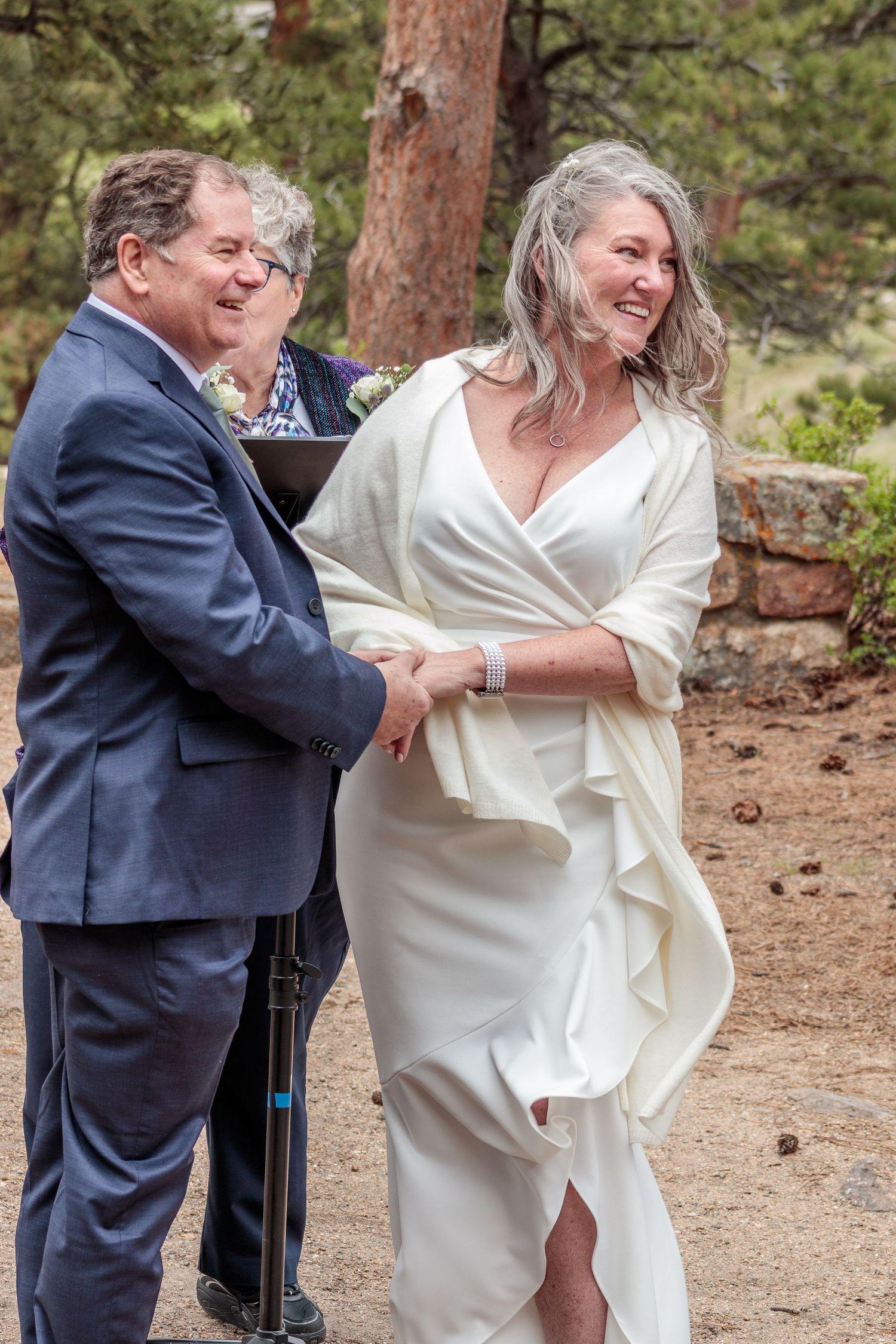 A couple in wedding attire dances playfully in a forest setting with pine trees in the background.