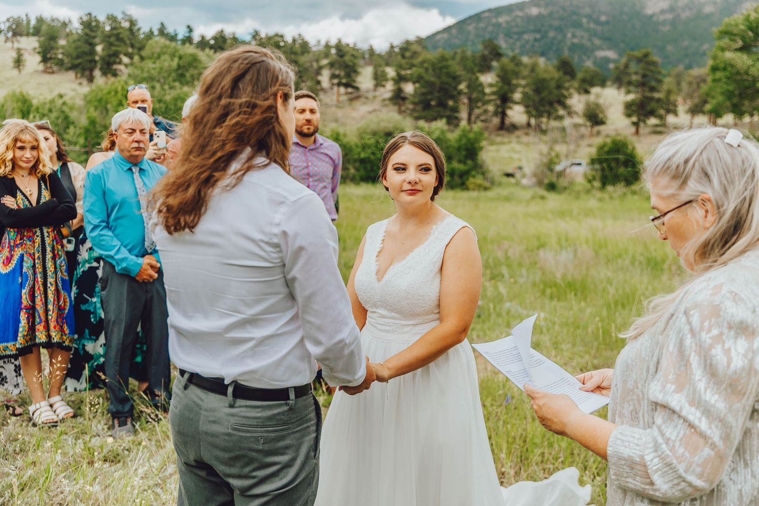 A group celebrates at an outdoor wedding ceremony in a scenic mountain meadow with green grass and trees.