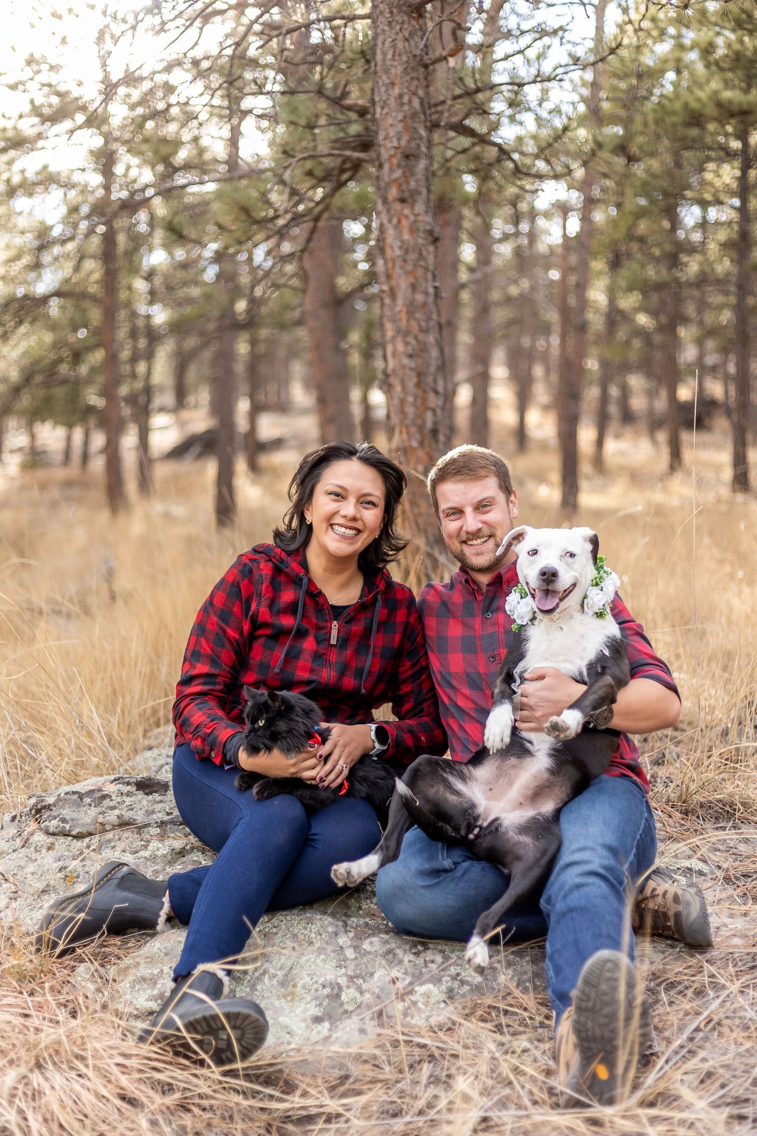 A couple in matching buffalo plaid shirts sits on the ground with their two dogs in a rustic outdoor setting.