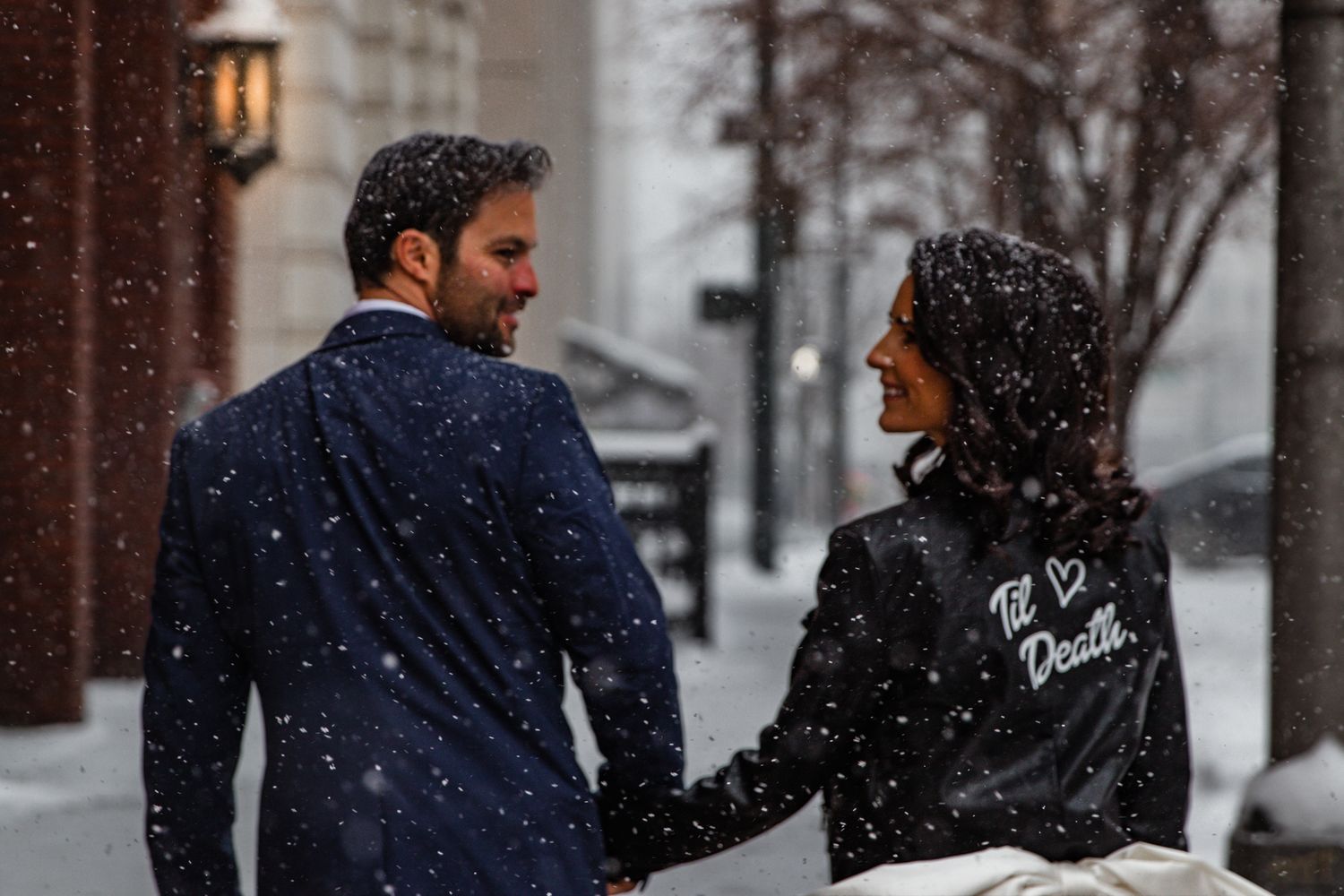 People walking through a snowy city street during winter with snowflakes falling and buildings in the background.