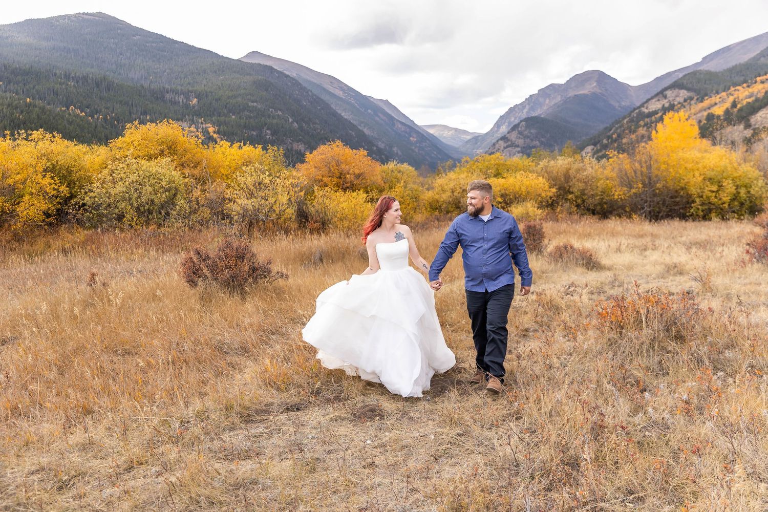 A couple walks hand in hand through a golden autumn meadow with mountains and yellow aspen trees in the background.