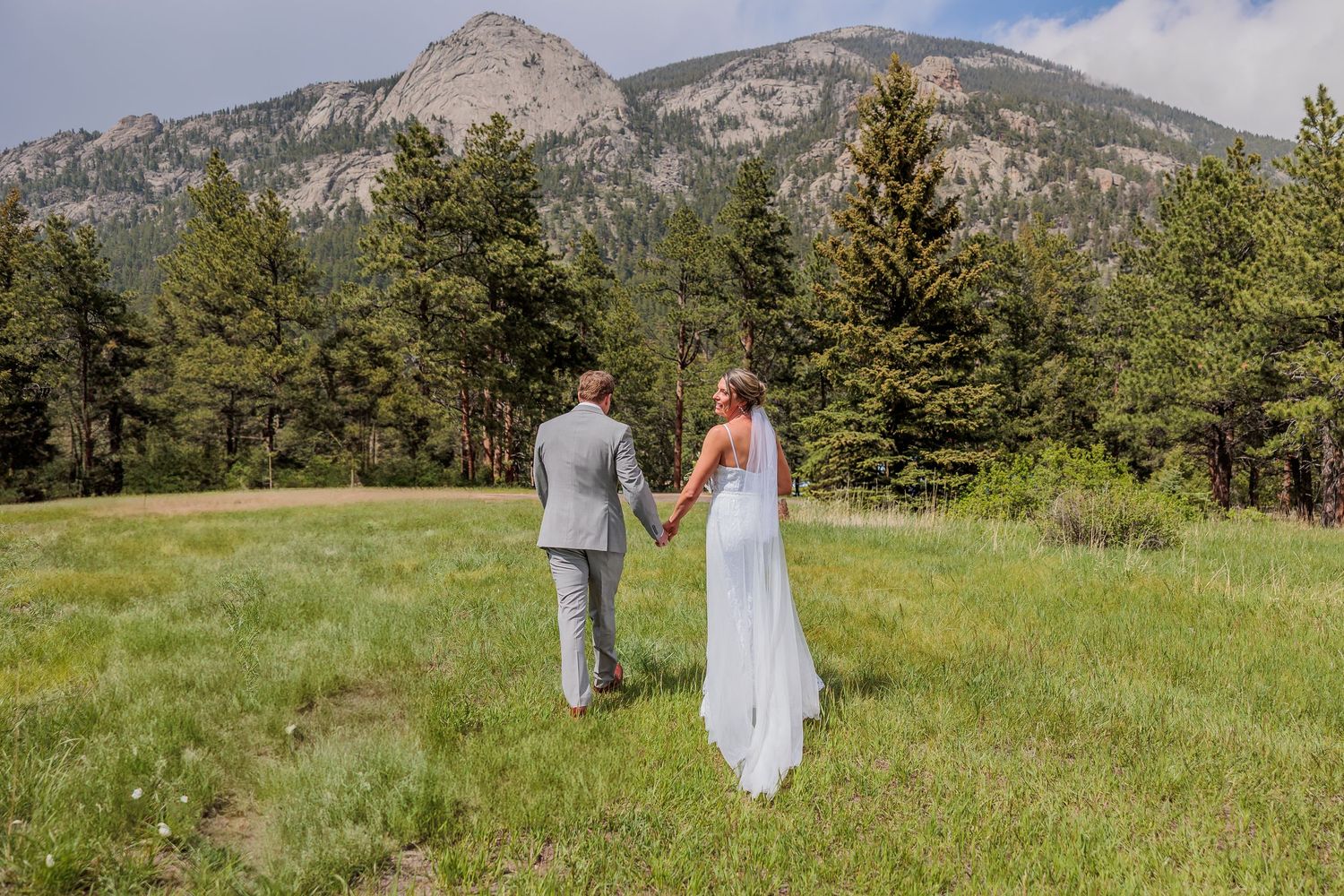 A couple walks through scenic mountain meadows with evergreen trees and rocky peaks in the background.