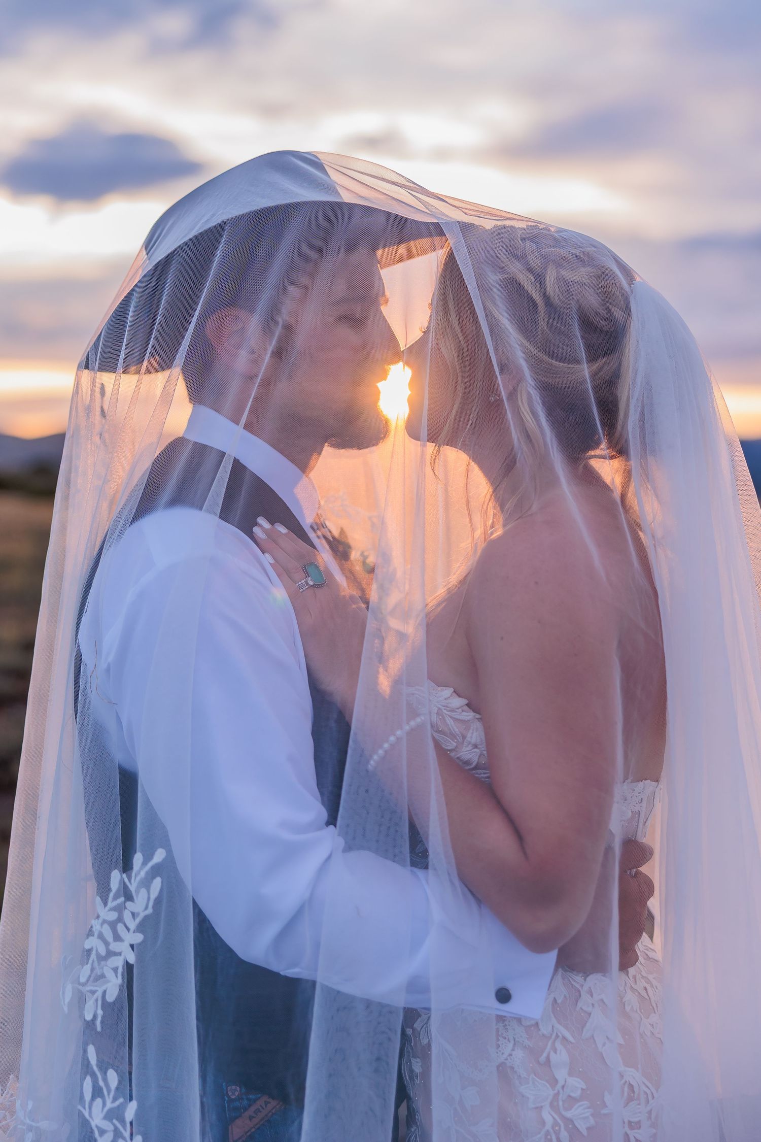 A romantic wedding couple shares tender moments under a flowing veil during sunset.