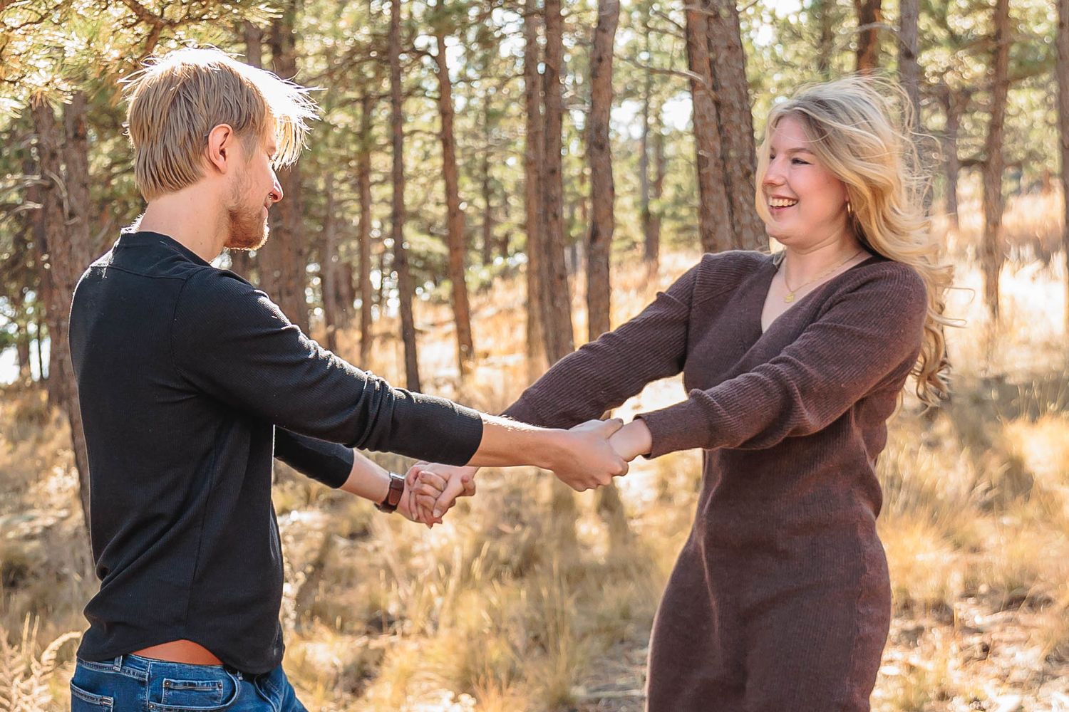 A couple in brown and black outfits hold hands while walking through a sunlit pine forest during autumn.