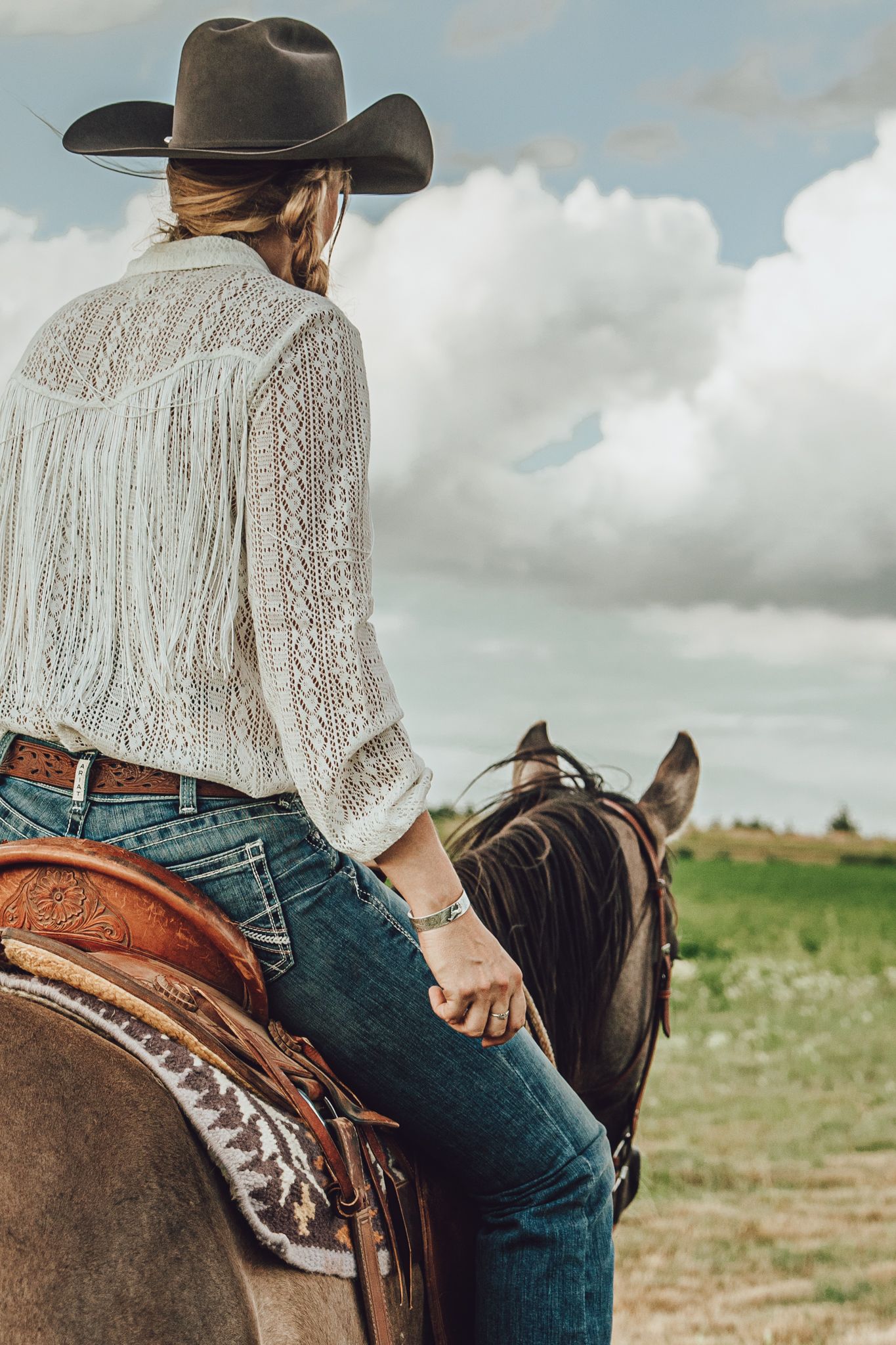 A cowgirl in a lace top and jeans sits on horseback against a cloudy prairie landscape.