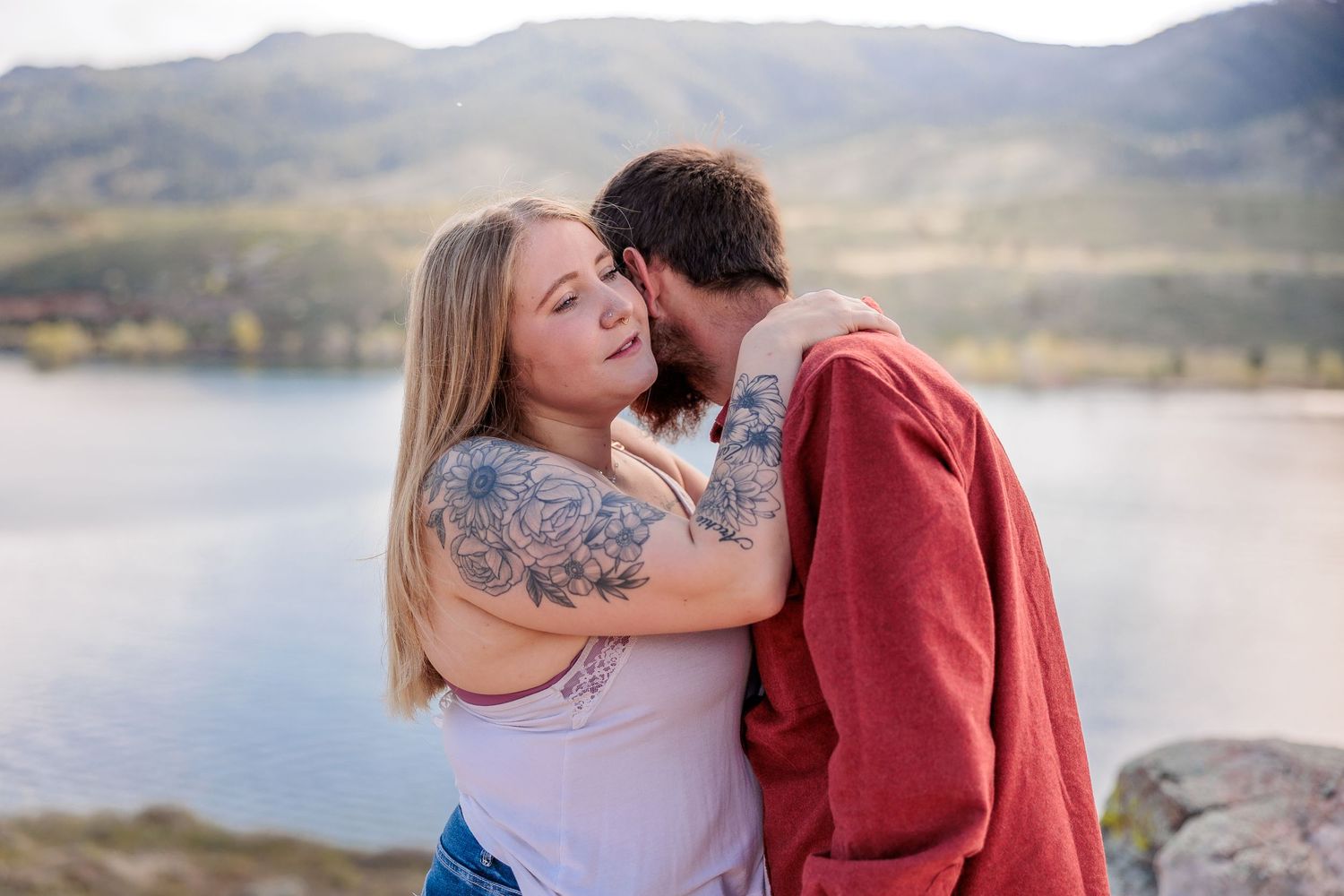 A couple shares romantic moments by a serene lake with mountains in the background during golden hour.