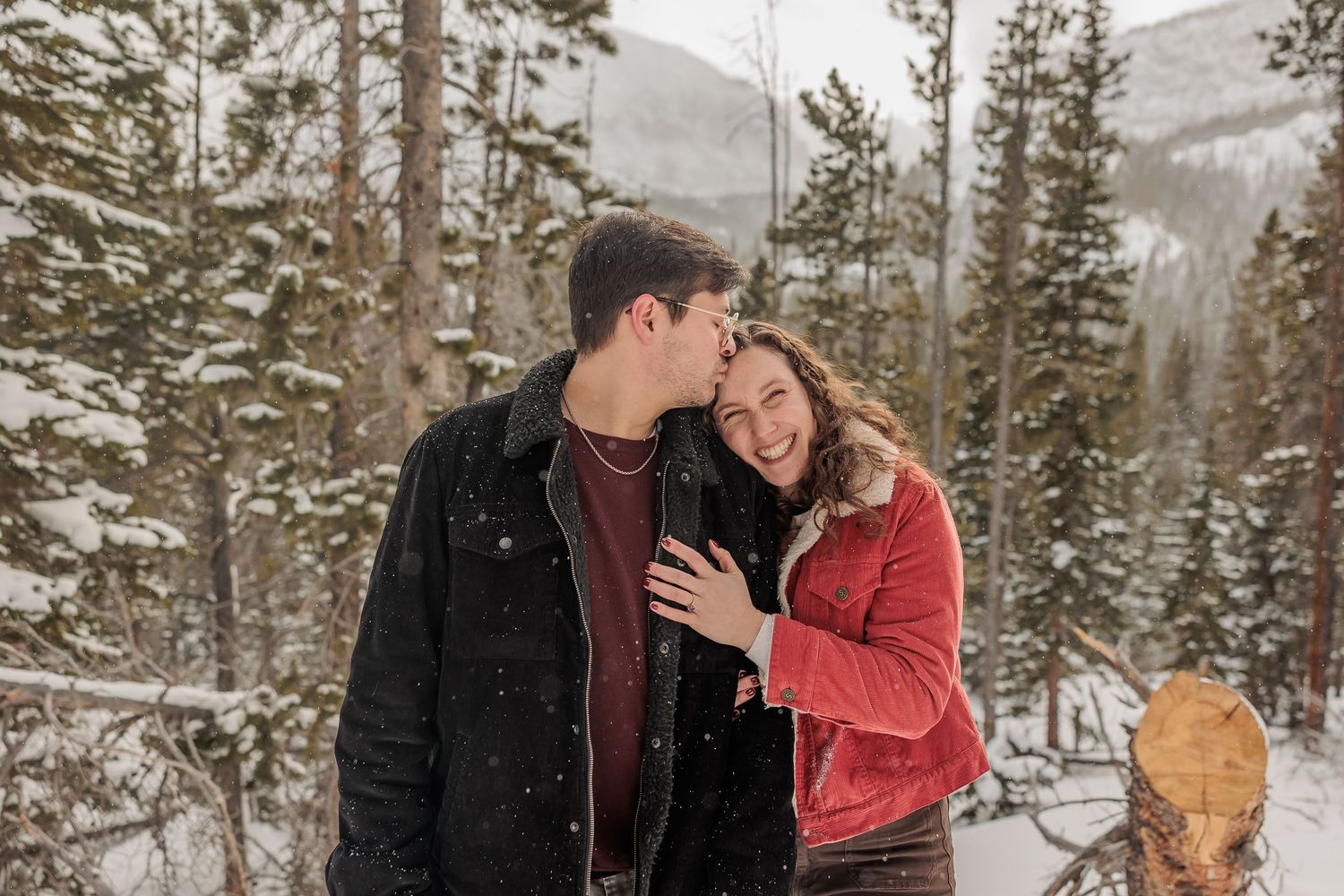 A couple shares an intimate moment in a snowy mountain forest setting wearing winter coats.