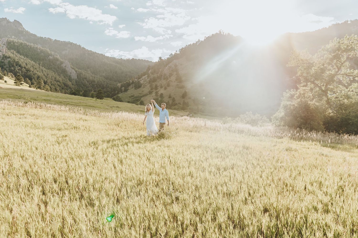 Two people walking hand in hand through a golden wheat field with mountains and sunlight in the background.