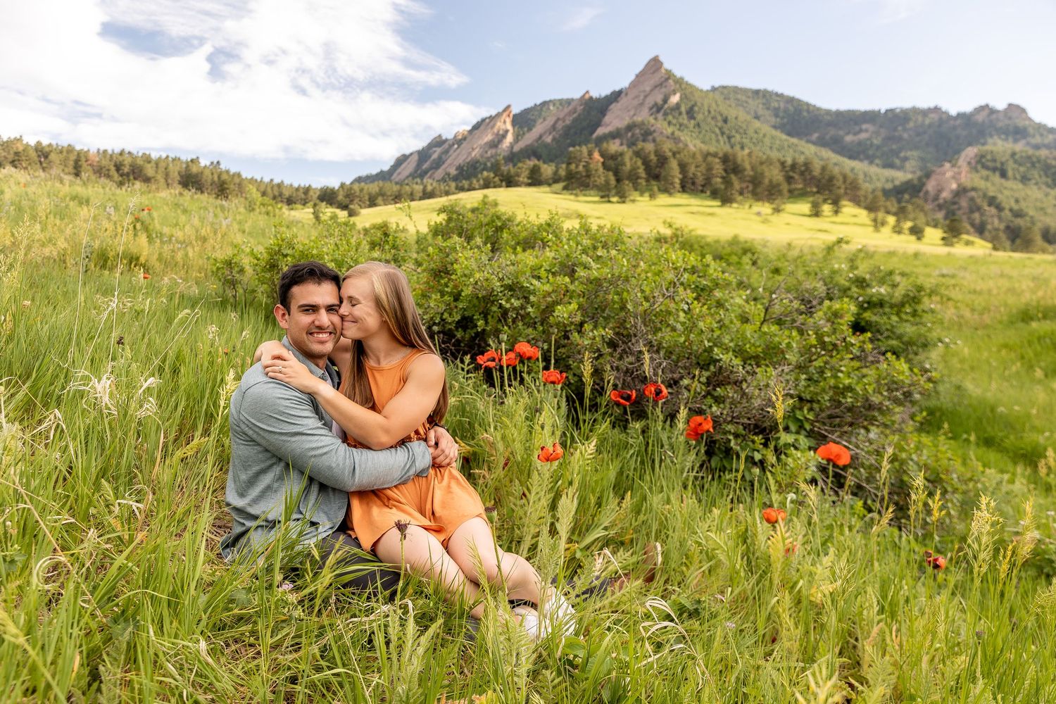 A couple sits together in a grassy meadow with the Flatirons mountains and red wildflowers in Boulder, Colorado.