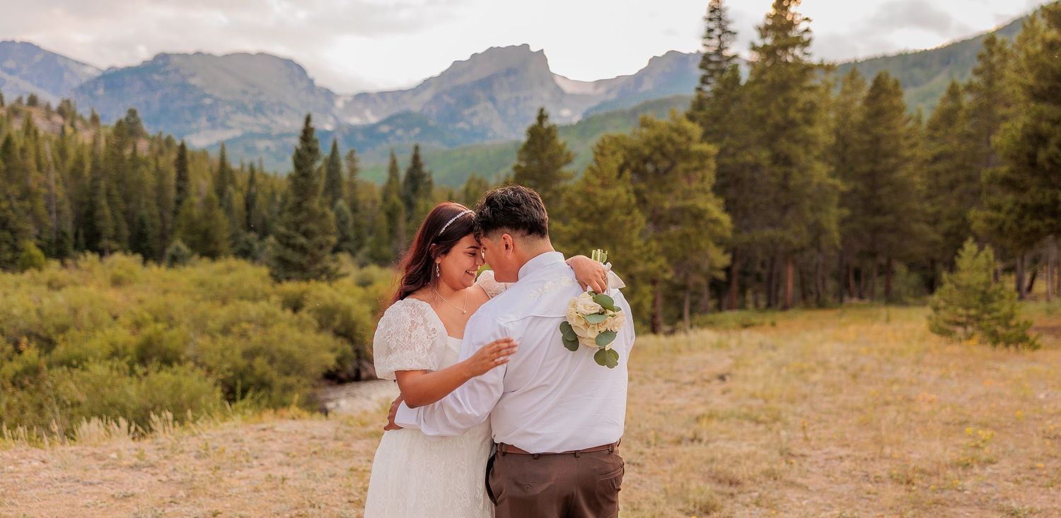 A couple embraces in a scenic mountain meadow surrounded by evergreen forest and peaks.