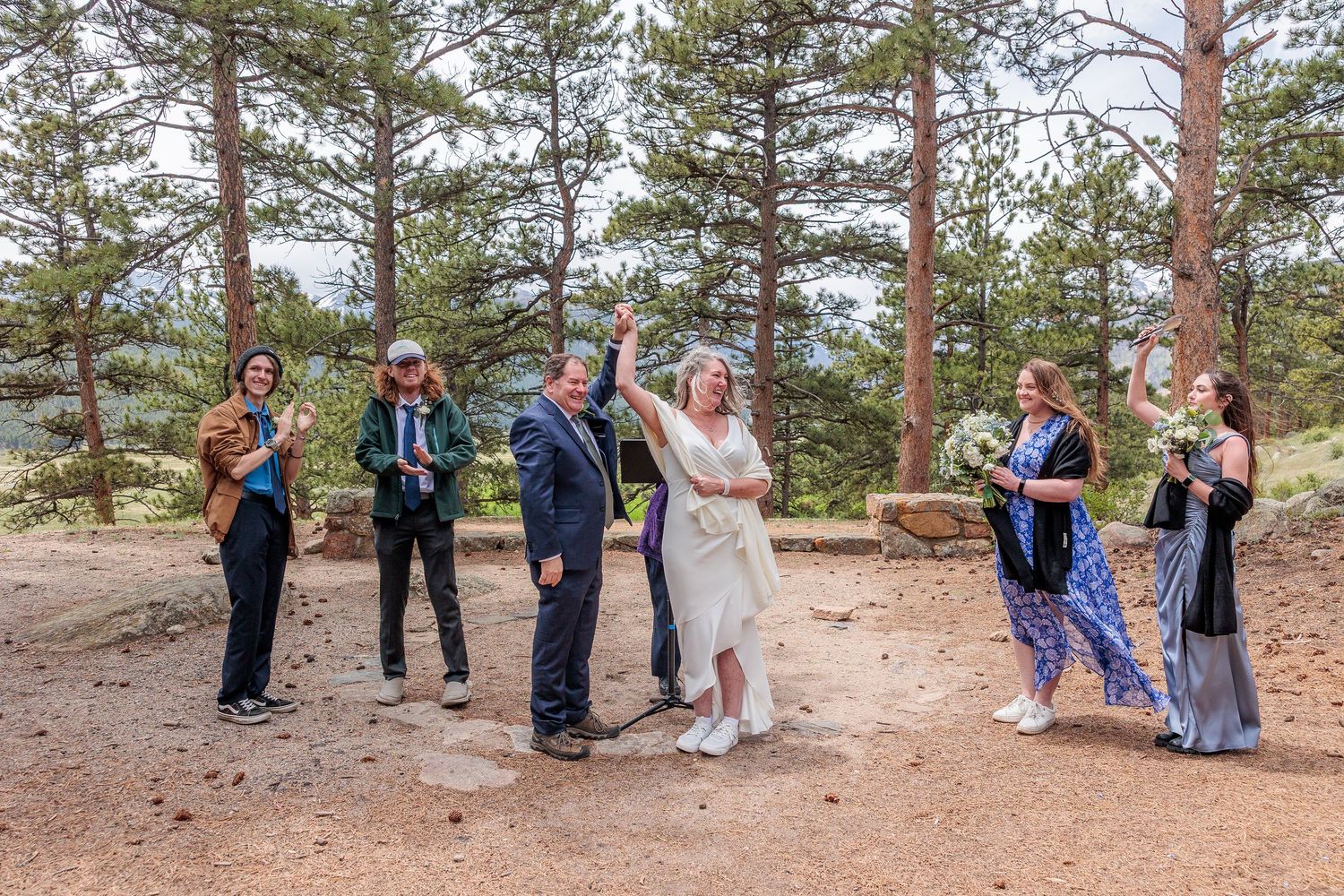 Wedding party poses beneath tall pine trees during an outdoor mountain ceremony.