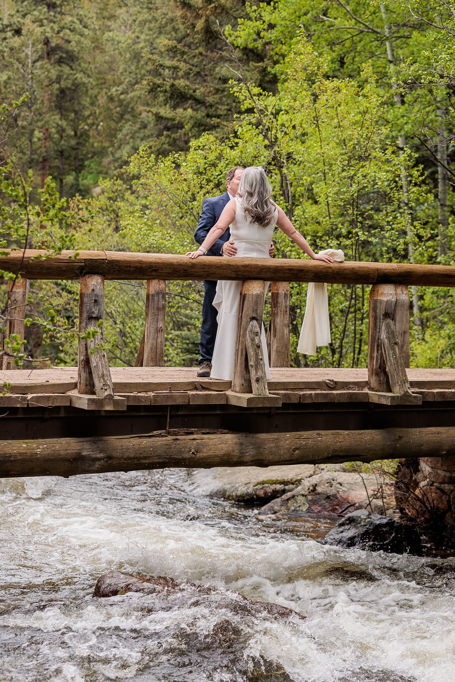 Couple standing on a rustic wooden bridge over rushing whitewater rapids in a lush forest setting.