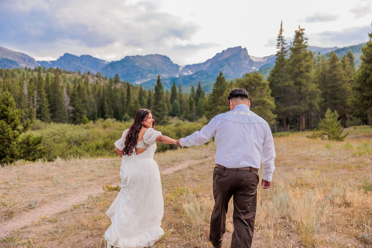 Couple walking hand in hand through scenic mountain meadow with evergreen forest and dramatic peaks in background in Rocky Mountain National