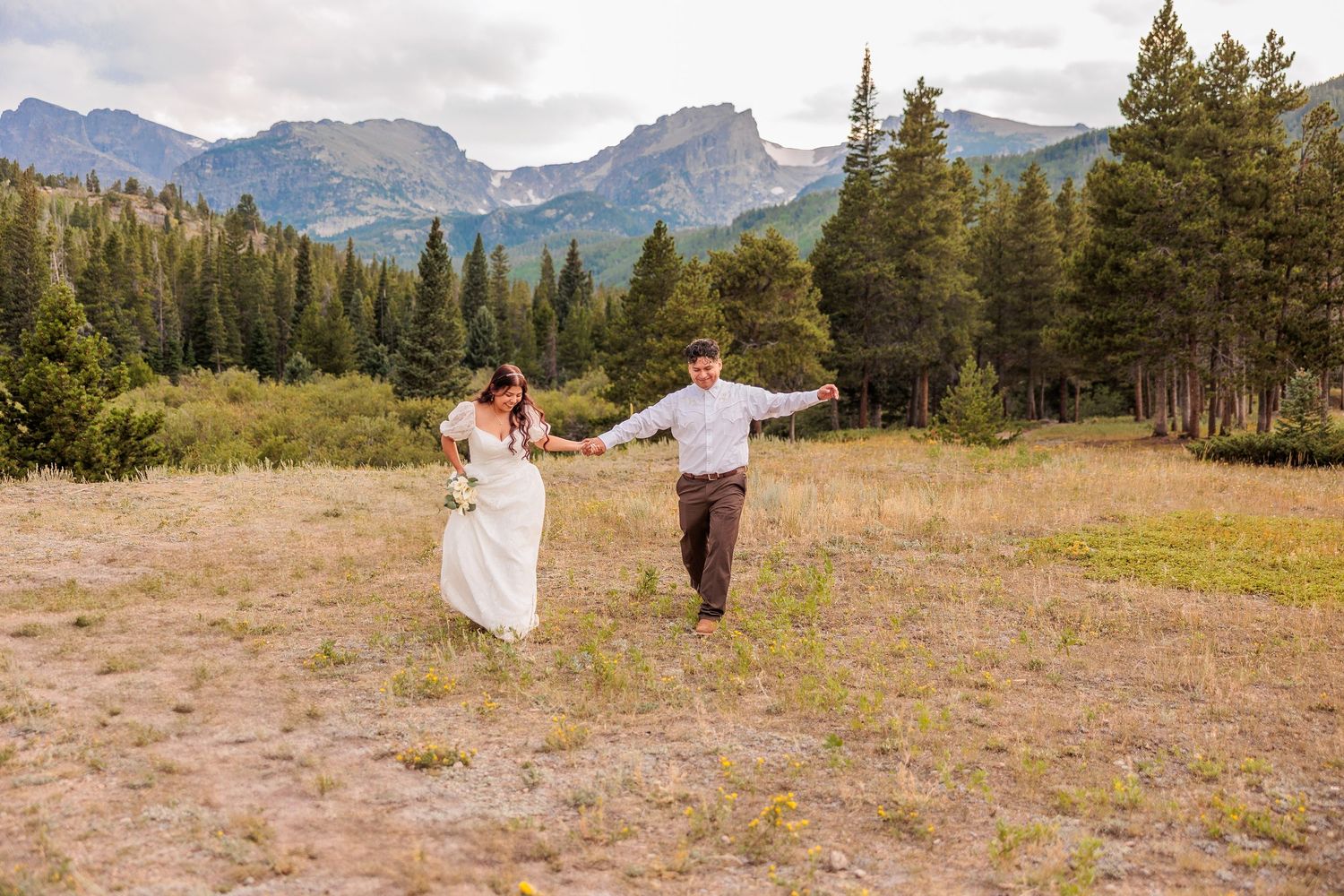 A couple celebrates their wedding day in a scenic mountain meadow surrounded by evergreen trees and distant peaks.