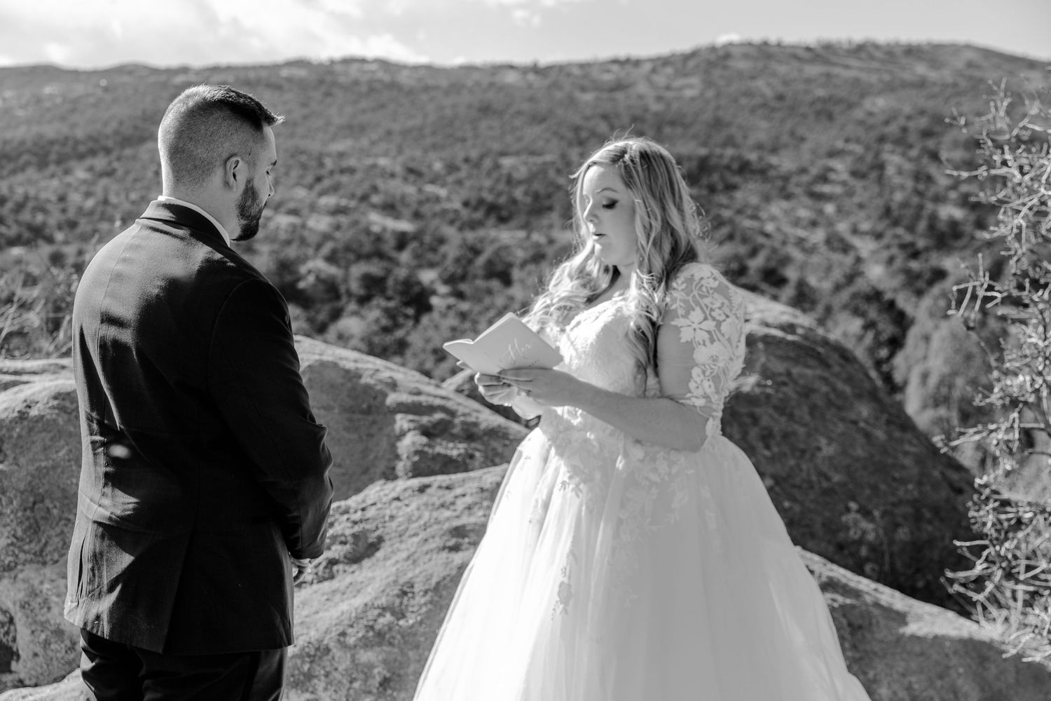 A couple in wedding attire stands on a scenic mountain overlook with sweeping valley views in the background.