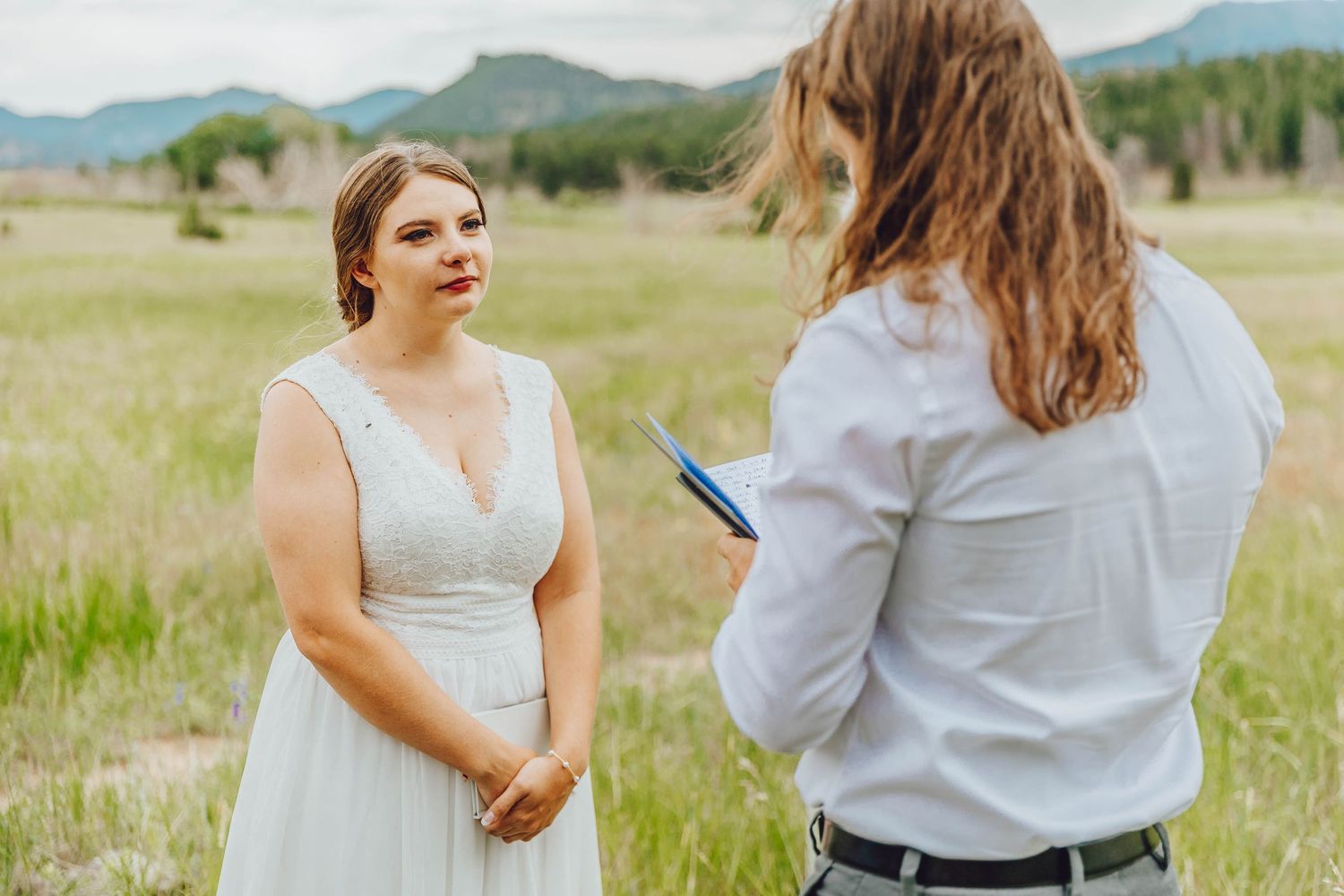 Two people in coordinating gray and white outfits stand together in a grassy field with mountains in the background.