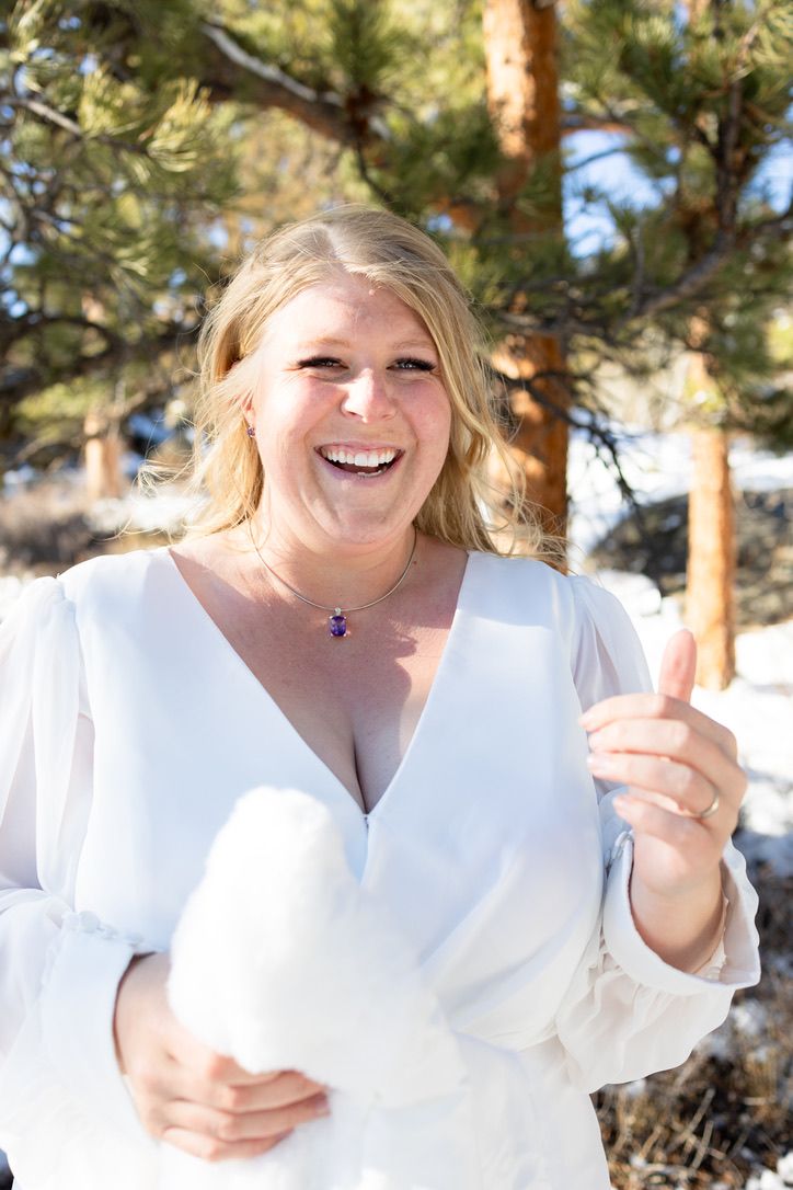 Smiling blonde bride in white dress giving thumbs up outdoors in snowy winter setting with pine trees.