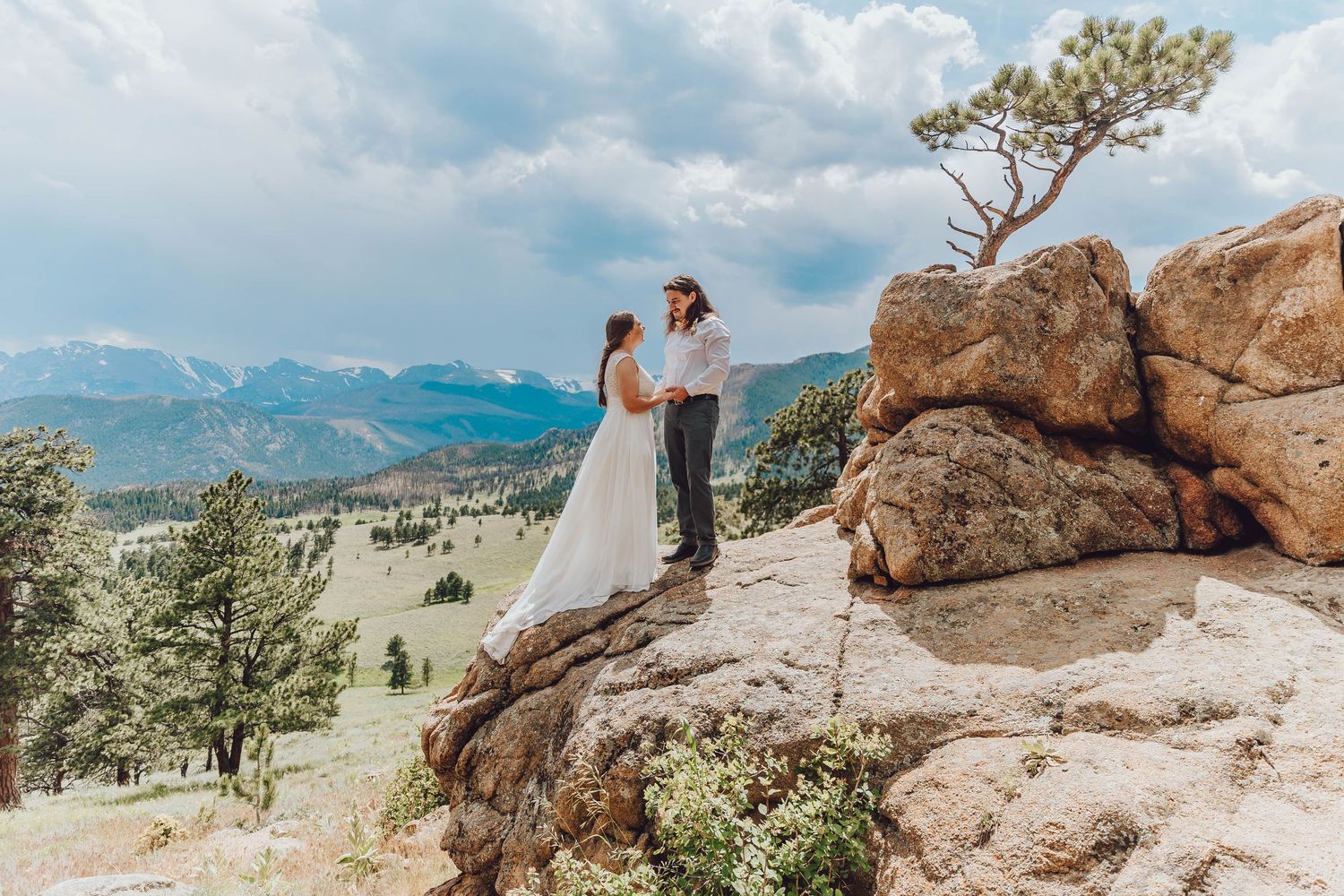 A couple in wedding attire stands on a scenic mountain overlook with dramatic rocky cliffs and evergreen trees.