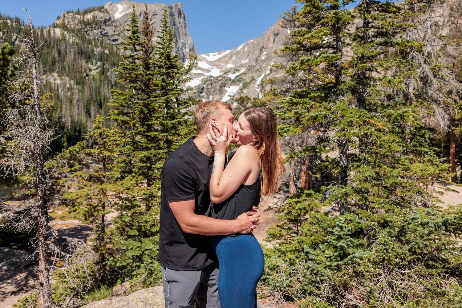 A couple shares a romantic moment surrounded by evergreen trees and rocky mountain peaks in a scenic wilderness setting.
