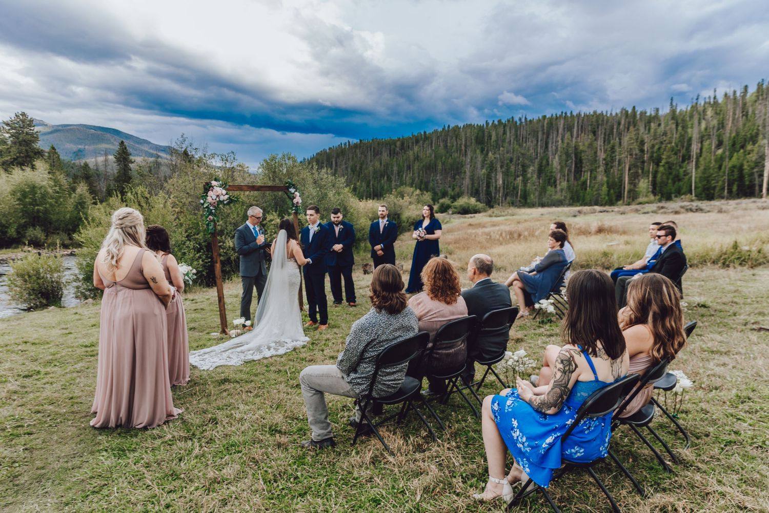 Wedding guests gather outdoors in a scenic mountain meadow setting with dramatic cloudy skies overhead.